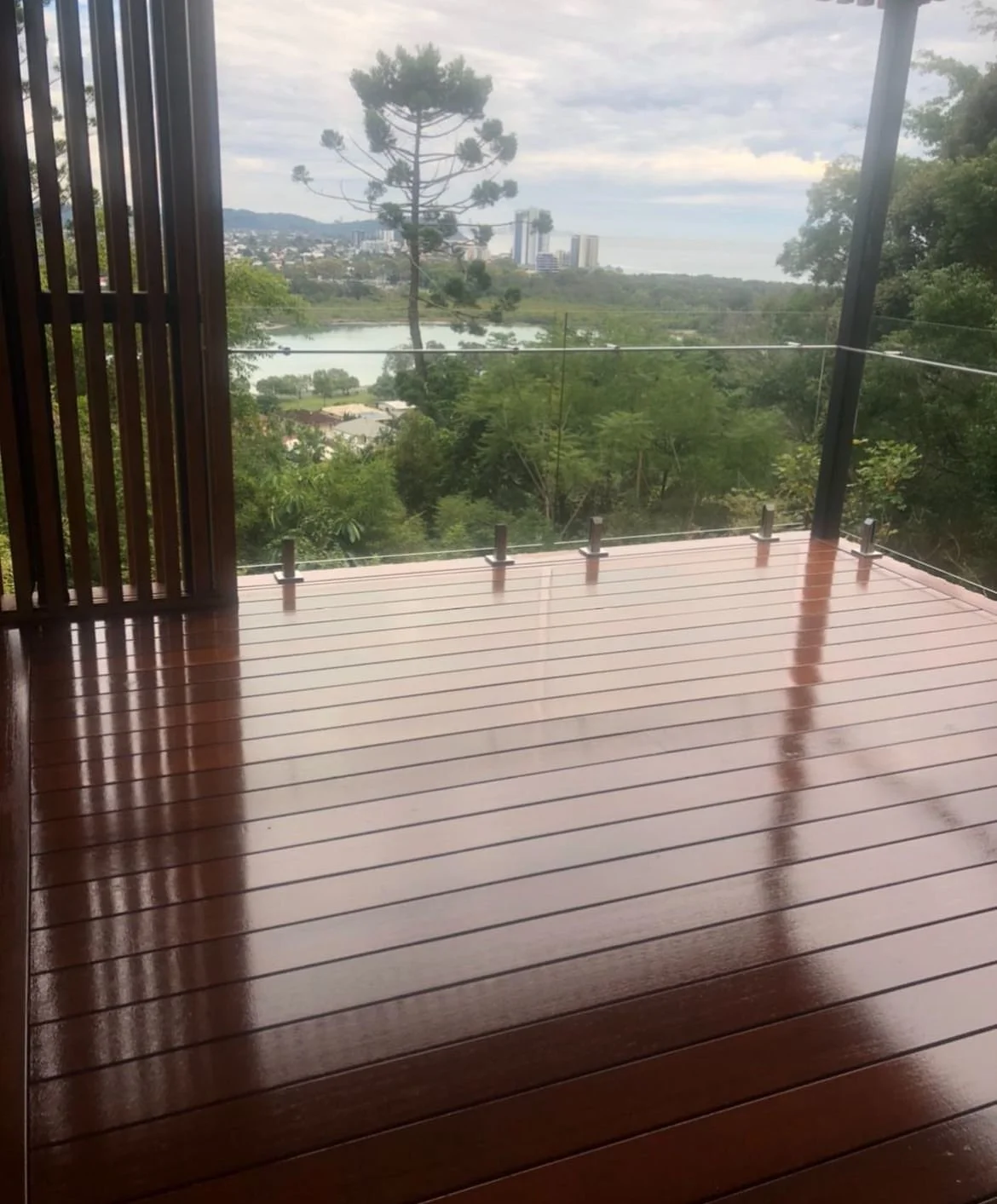 A wooden balcony with glass railing, overlooking a lush green landscape, a lake, trees, and a distant city skyline under a cloudy sky.