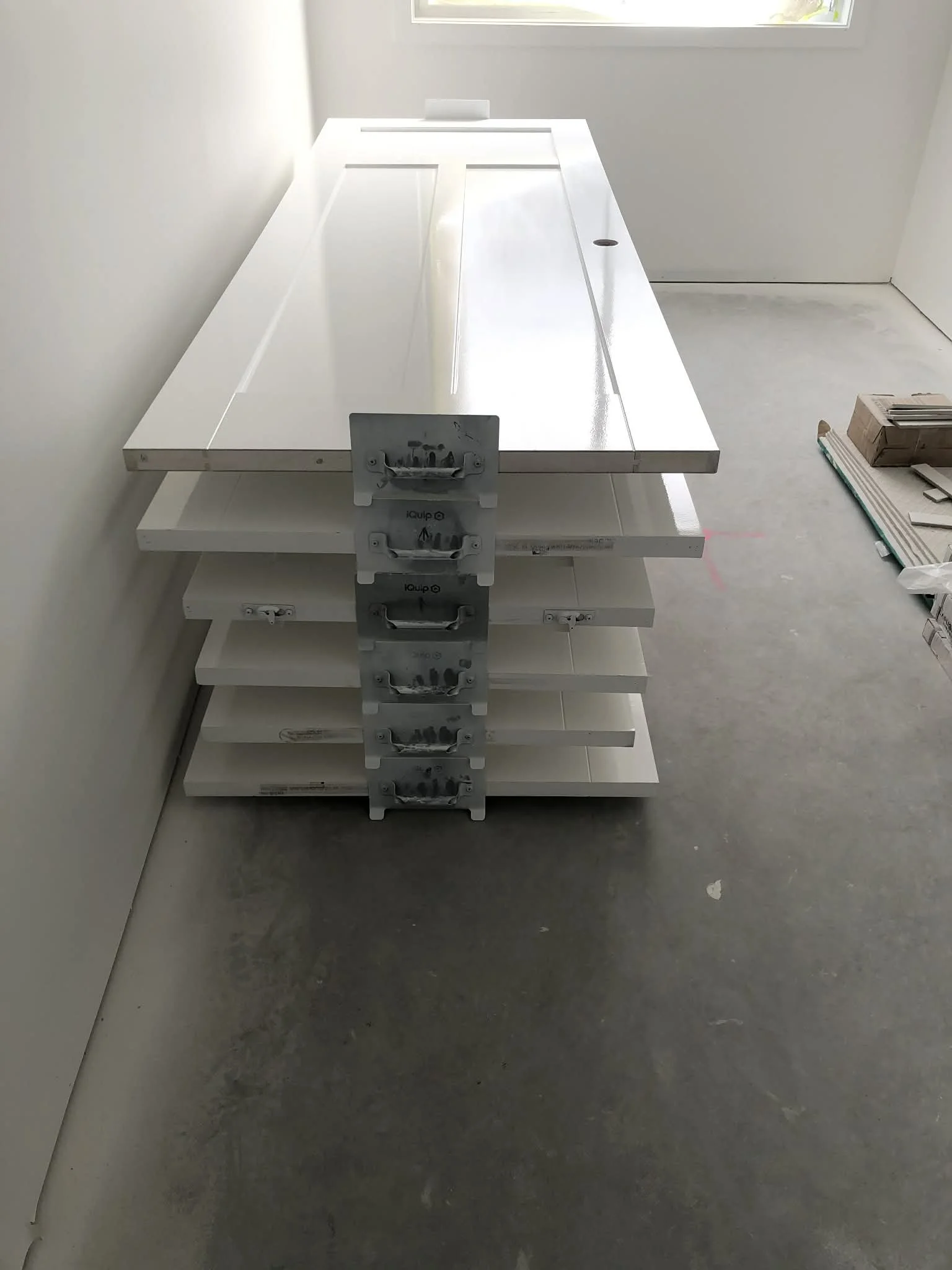 Stack of white cabinet panels and metal brackets in a room under construction, with construction materials on the floor and a window in the background.
