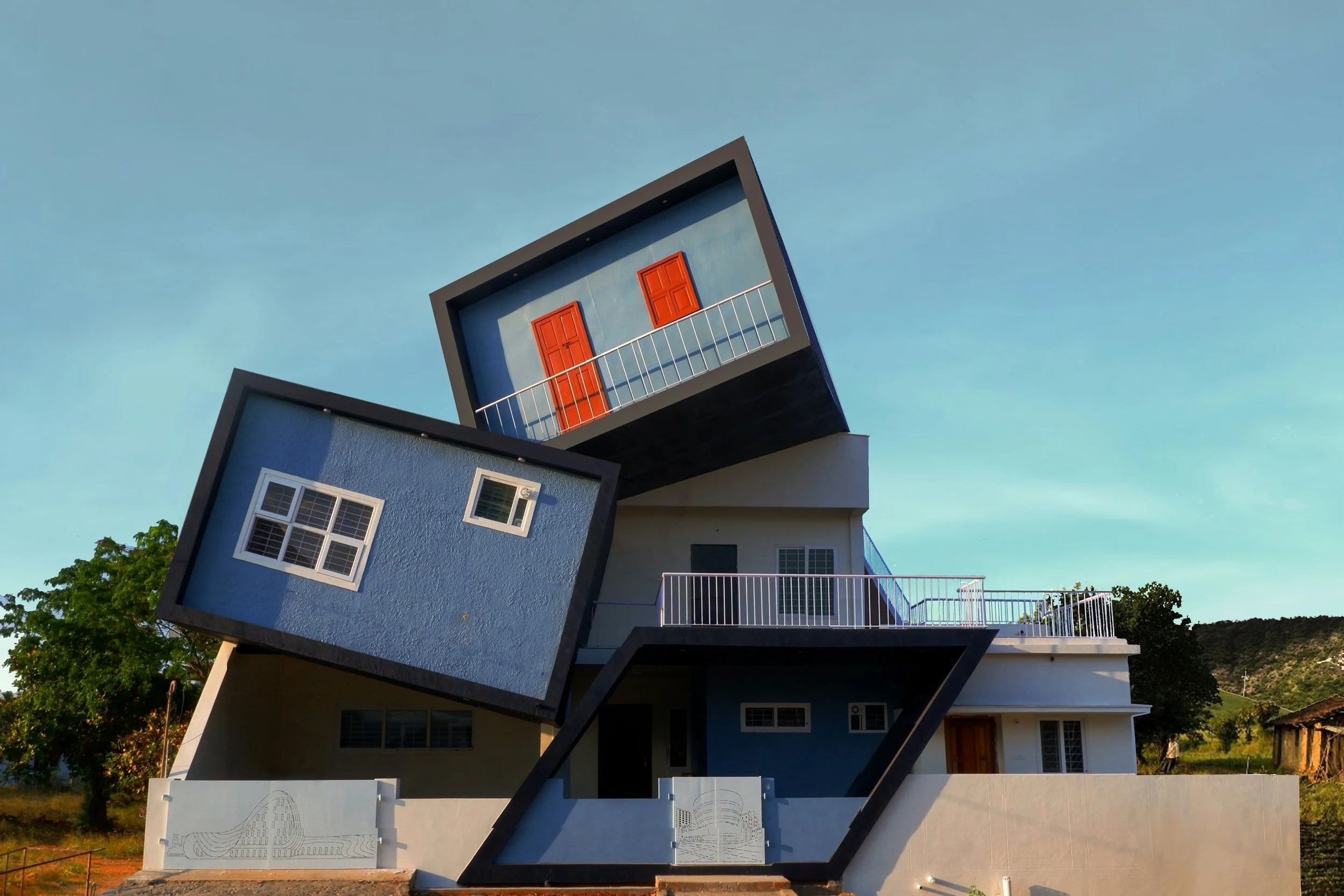 A modern building with a surreal architectural design featuring multiple cubic sections at different angles, with windows and balconies, set outdoors against a clear sky and distant hills.