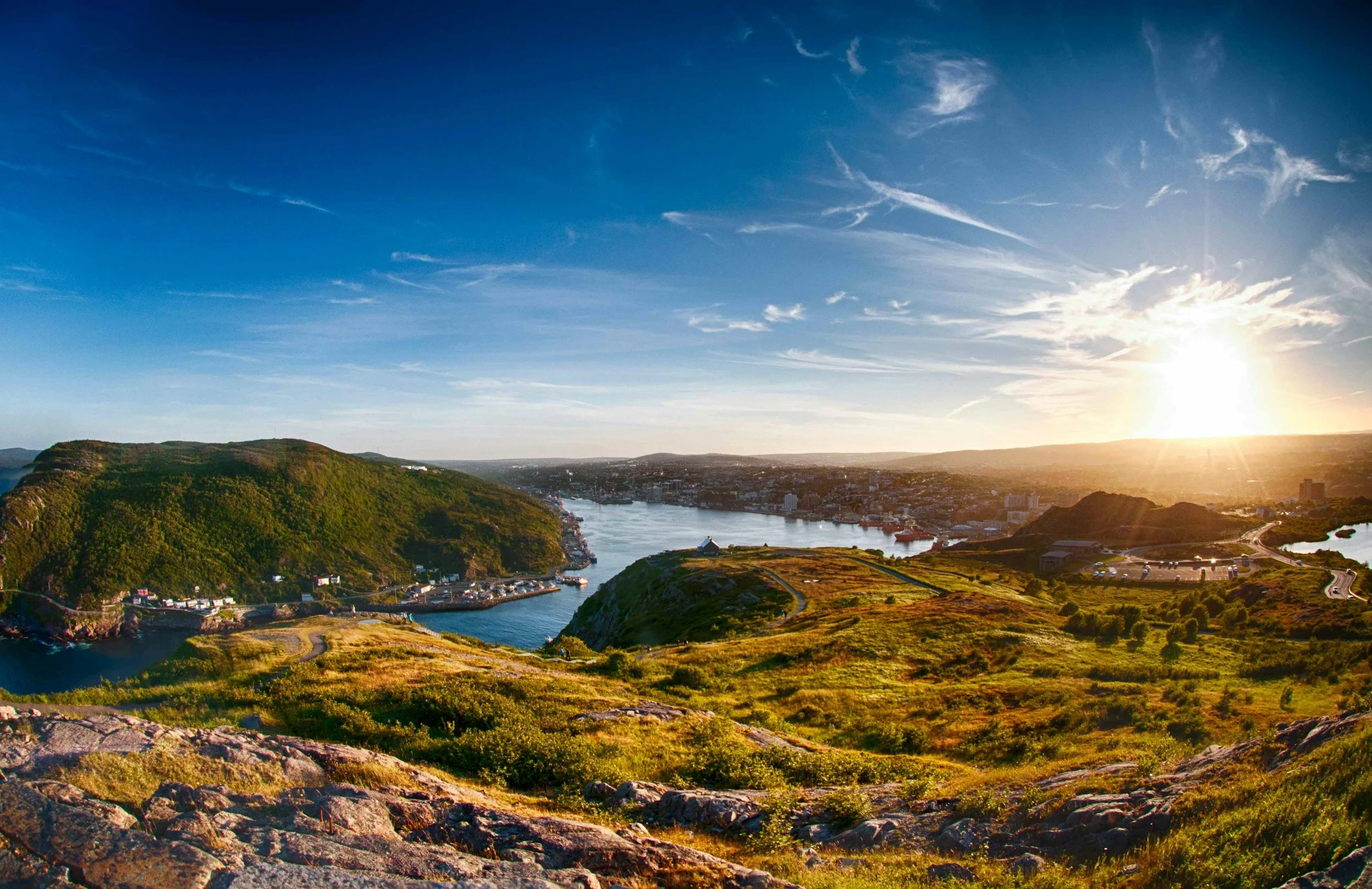 Sunset over a scenic coastal landscape with rolling green hills, a river, and a small harbor with boats.