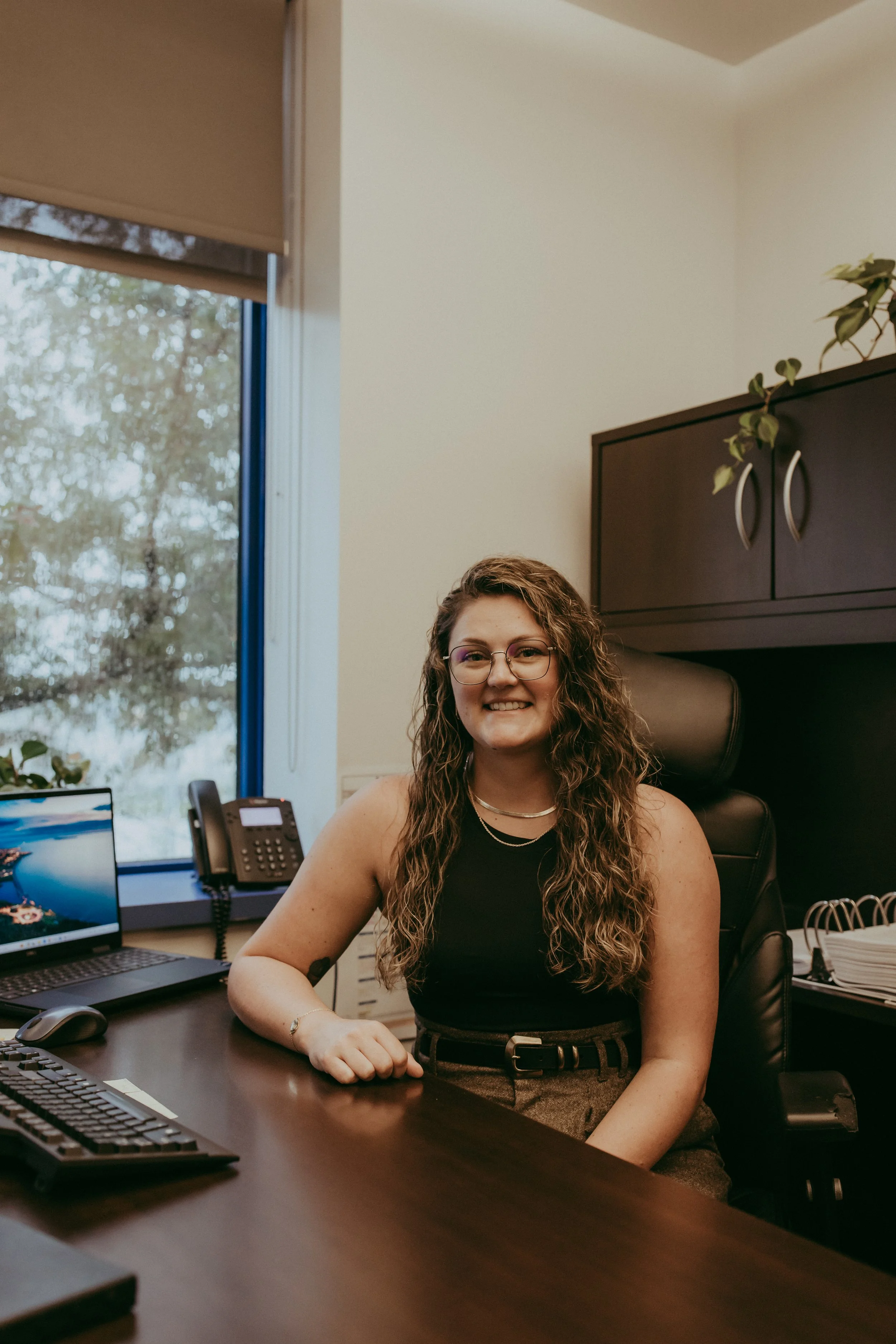 A woman with curly hair and glasses seated at a desk in an office, smiling at the camera.