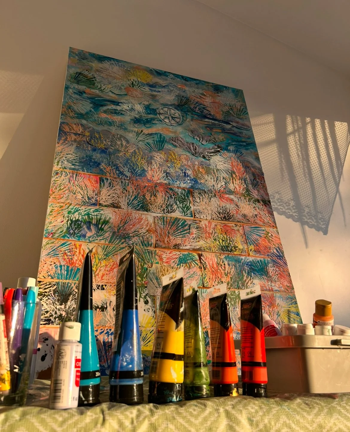 Tableau en cours de création avec peinture acrylique, pinceaux, et autres fournitures artistiques, avec un tableau coloré en arrière-plan.