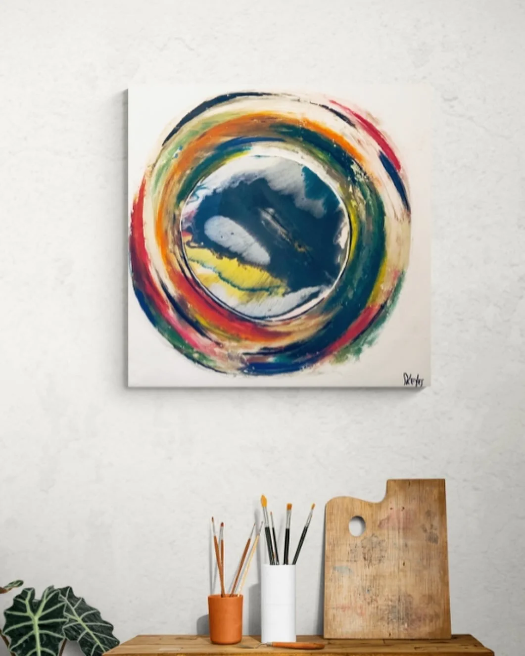 Une peinture abstraite avec un cercle coloré sur un fond blanc, exposée sur un mur. En dessous, une table avec des pinceaux dans des pots, une planche à découper en bois et une feuille de papier. Il y a aussi une plante en bas à gauche.