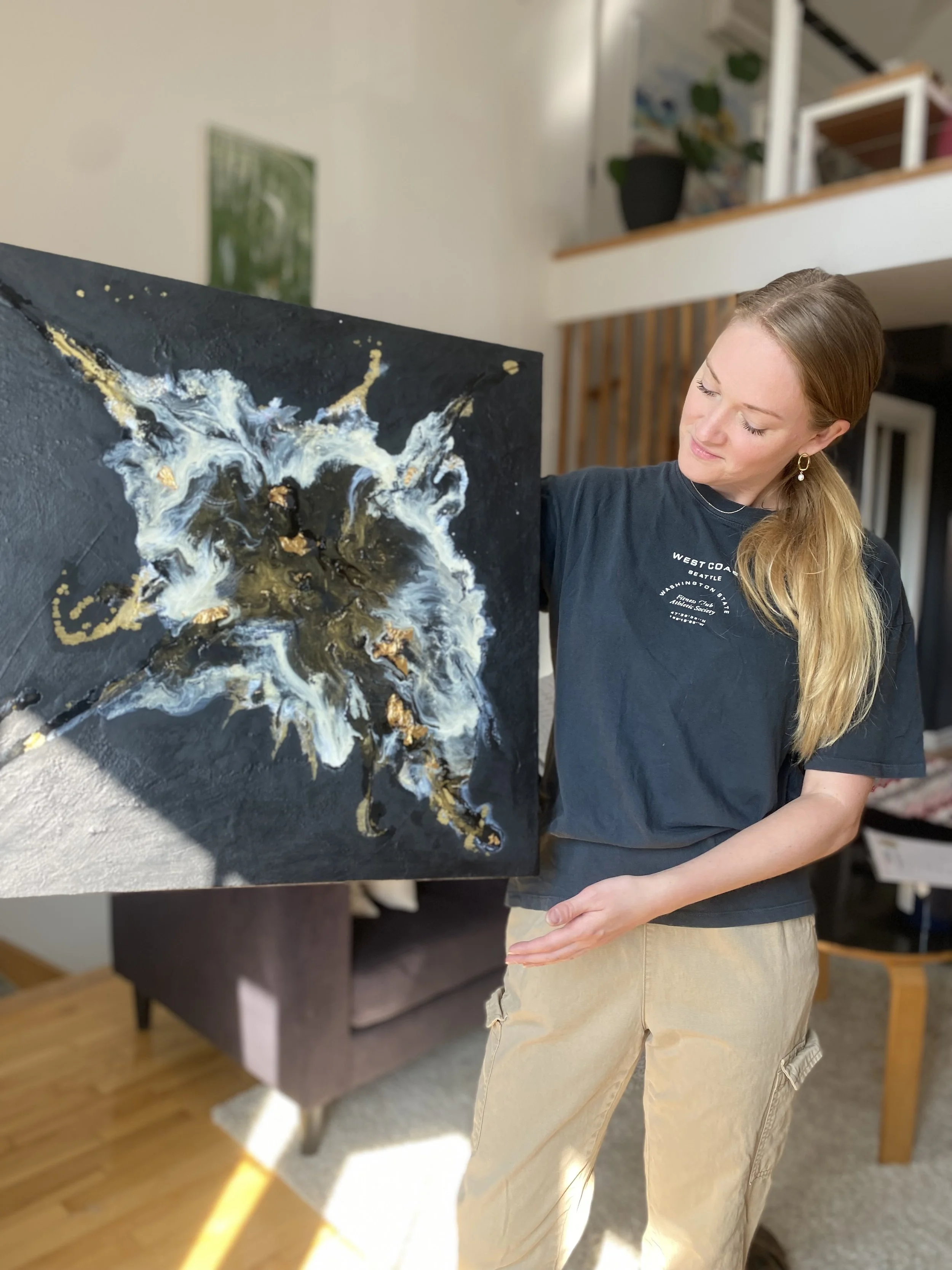 Une femme regarde une peinture abstraite. La peinture a des couleurs noir, blanc, or et blanc, avec des textures épaisses. Elle est dans un intérieur avec un canapé et des étagères en bois.