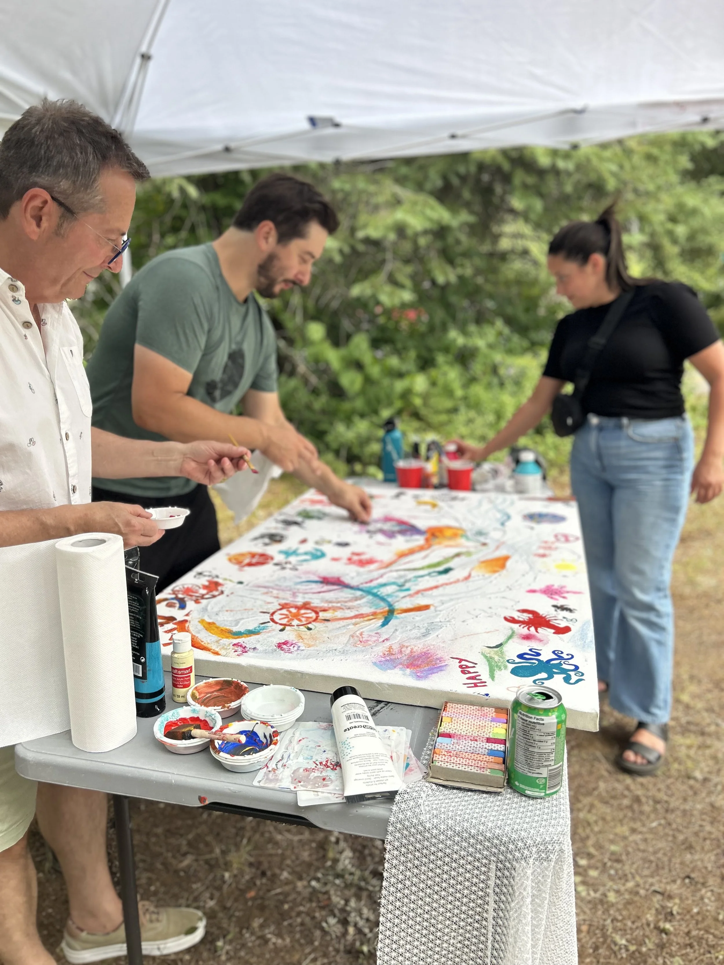 Groupe de trois personnes peignant une toile sous une tente en plein air, avec divers outils et peintures sur la table.