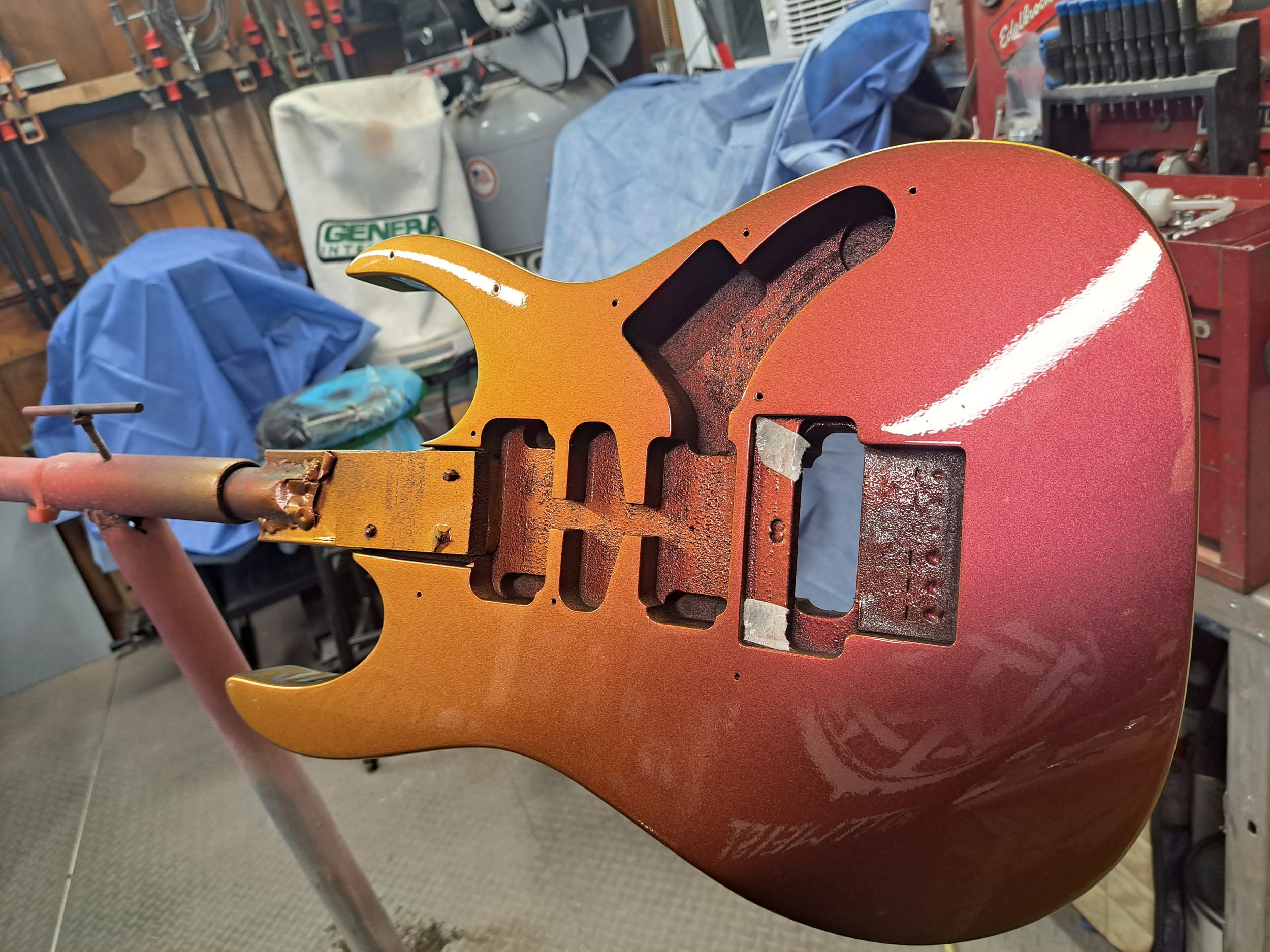 A guitar body in the process of being painted, with a gradient of red, orange, and yellow colors, mounted on a stand in a workshop.