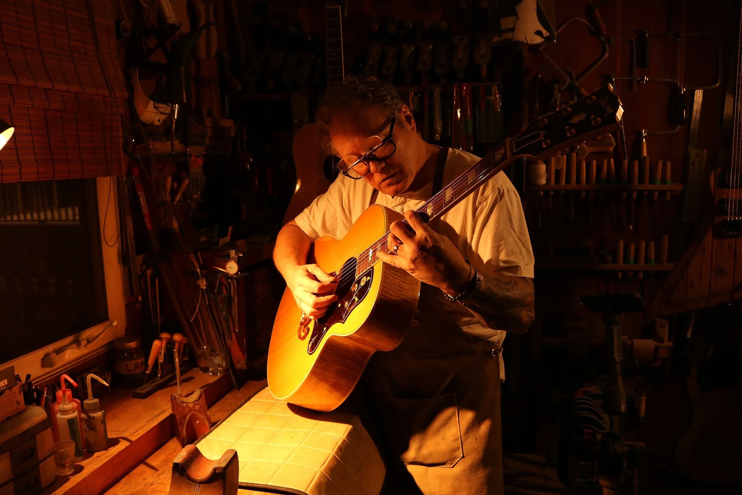 A man with glasses and tattoos playing an acoustic guitar in a dimly lit workshop or music studio.