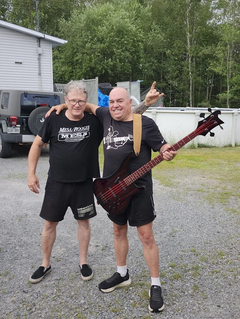 Two men smiling outdoors, one with his arm around the other, holding a bass guitar, in a backyard with gravel ground, trees, a house, and a white pool in the background.