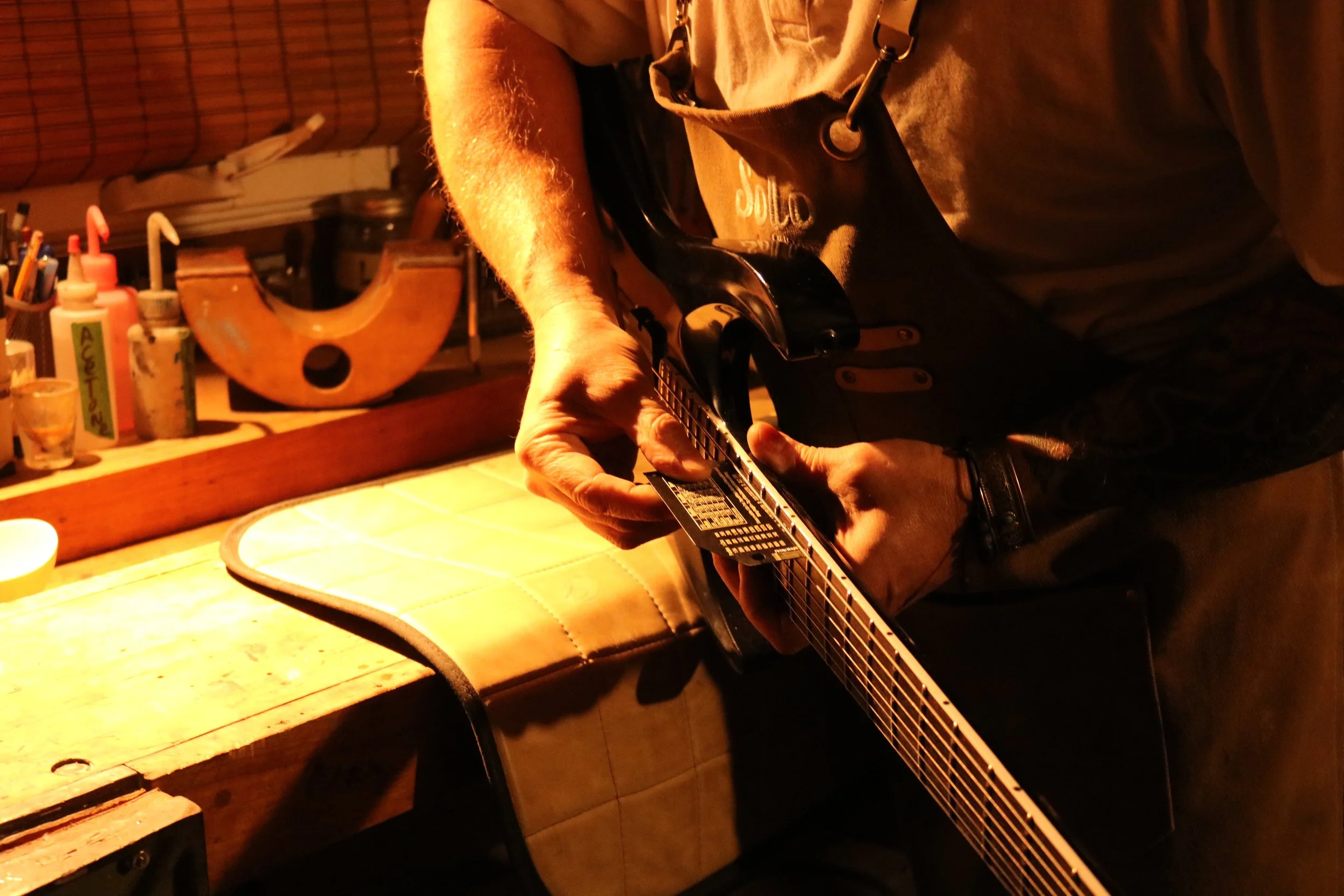 Close-up of a person playing an electric guitar in a dimly lit room with wooden workbench and various art supplies in the background.