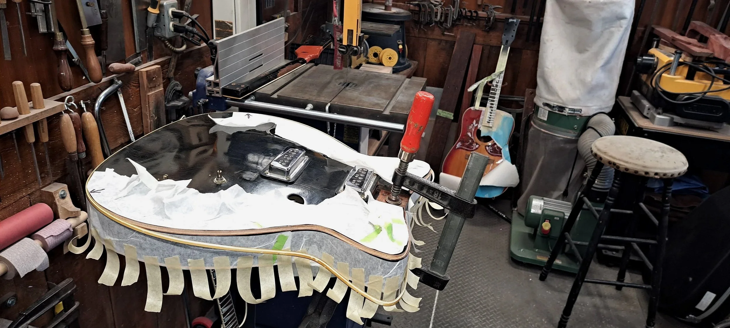 Workshop with an acoustic guitar on a workbench, surrounded by tools, clamps, and woodworking supplies.