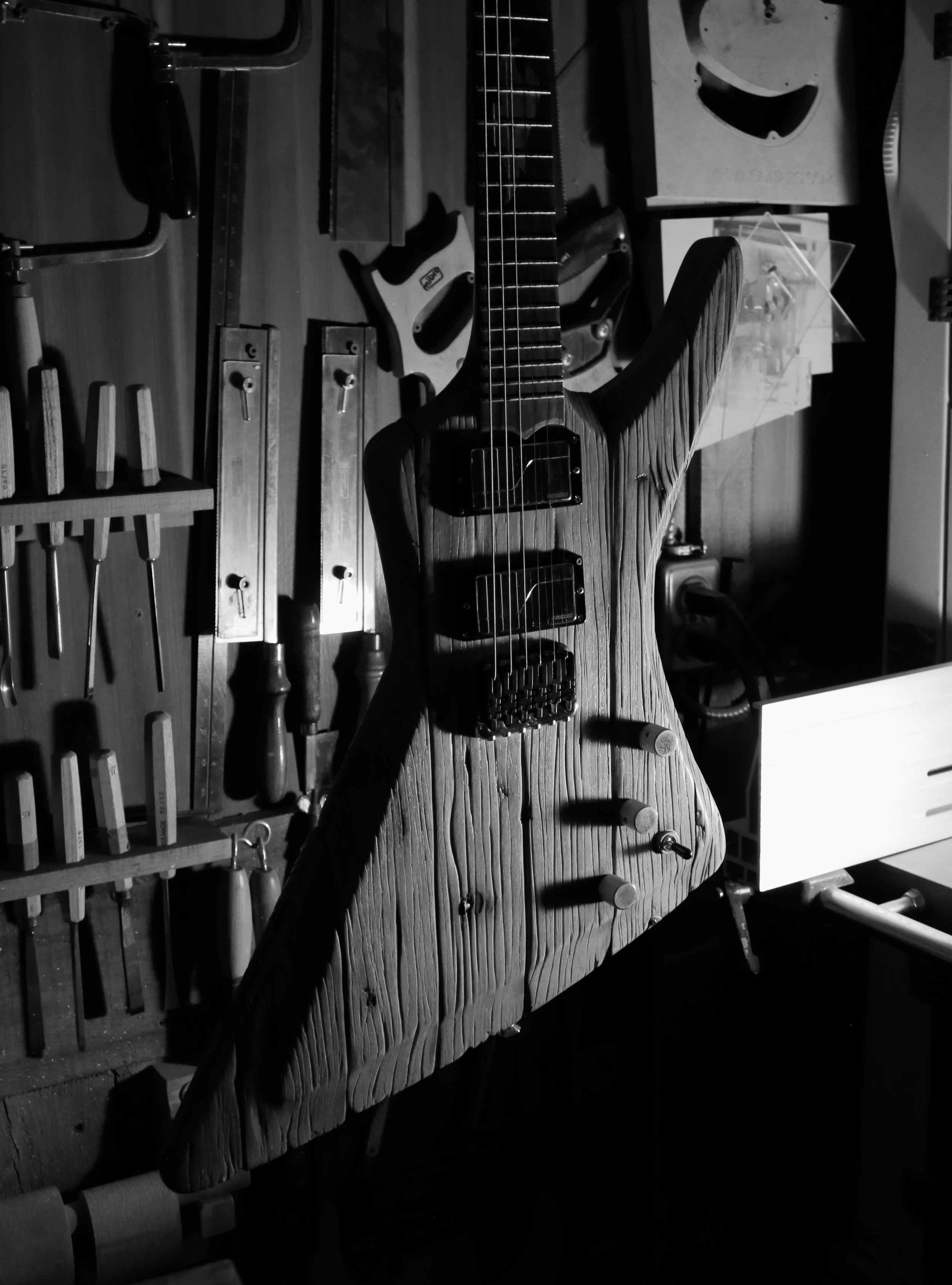 Black and white photo of an electric guitar with a wood grain texture, mounted on a workbench in a workshop, surrounded by woodworking tools.