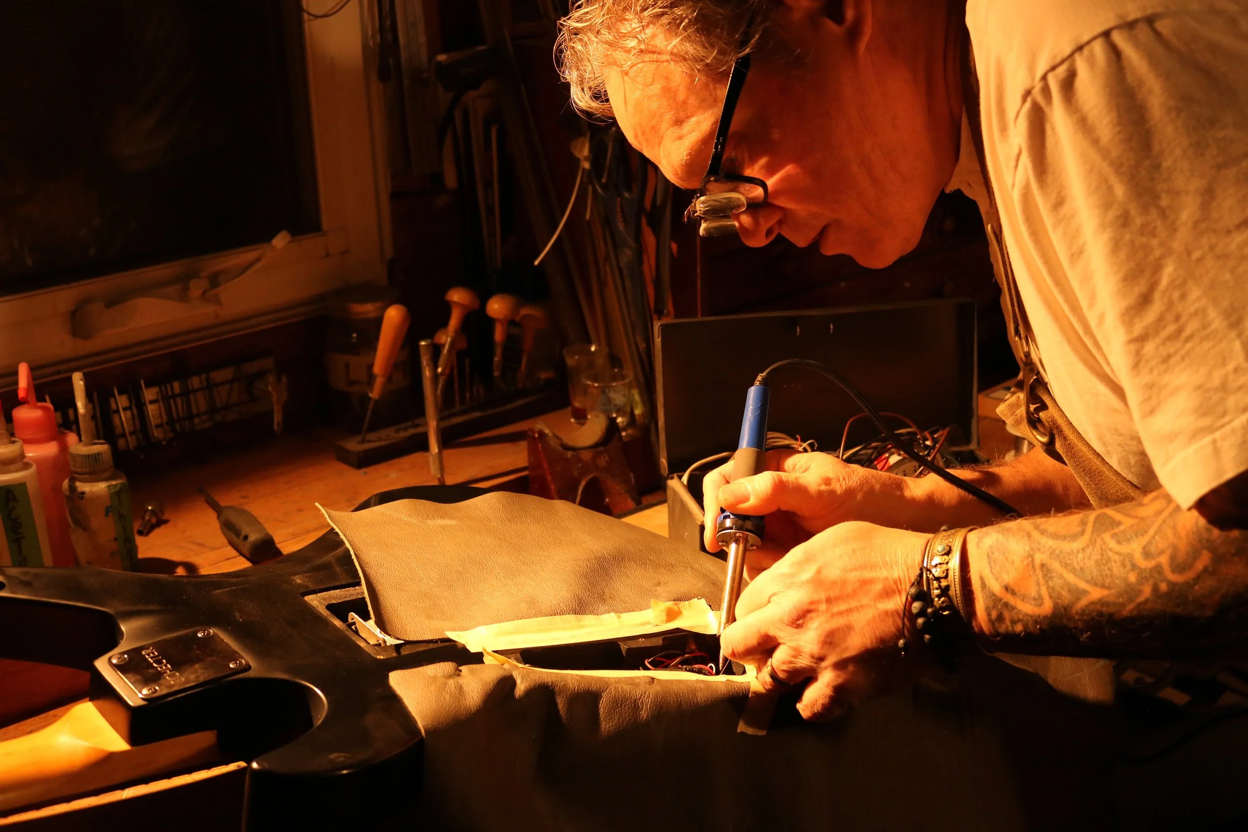 A person soldering a wire inside a black electric guitar body in a workshop with tools and supplies in the background.