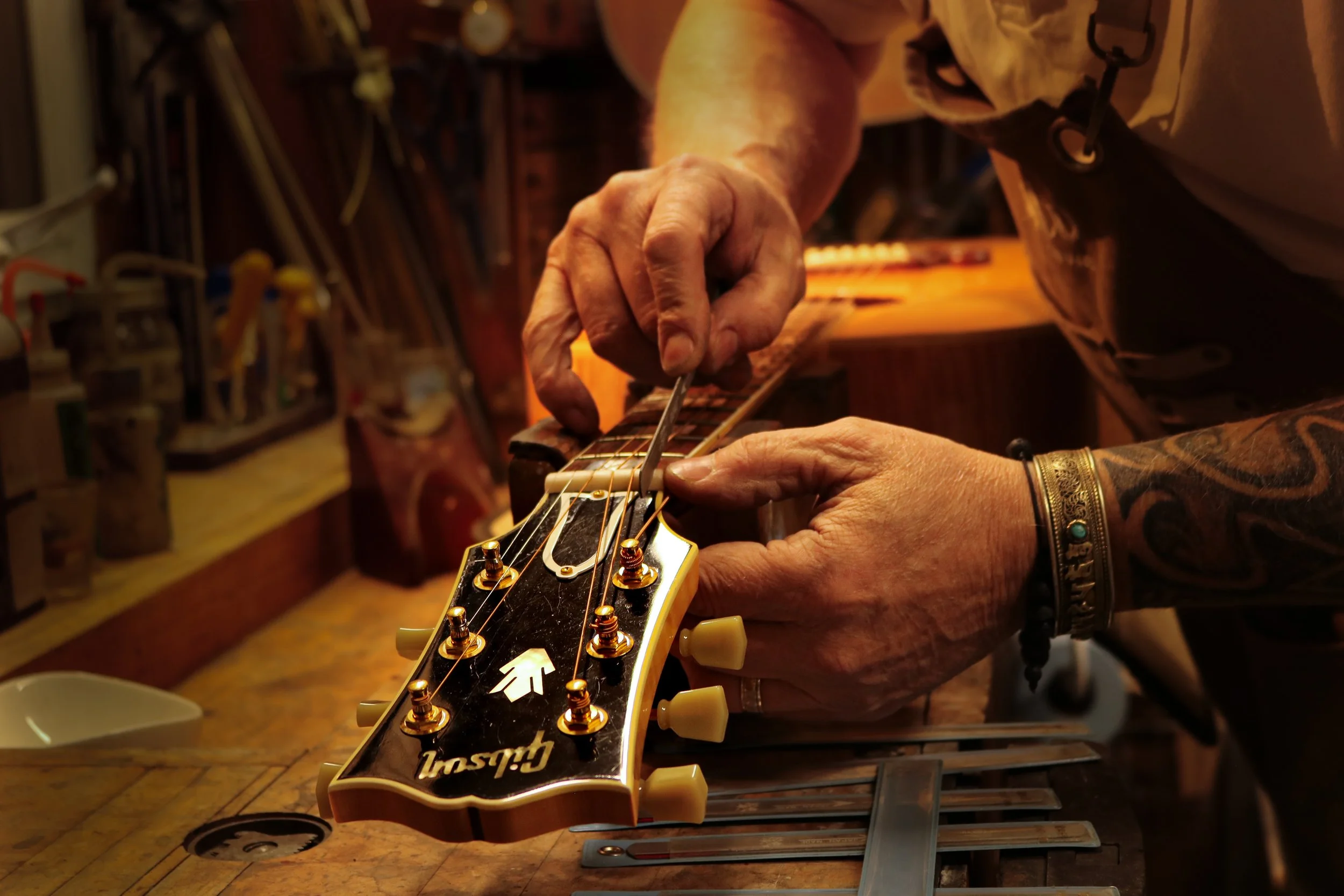 Close-up of a person making adjustments to the tuning of a Gibson electric guitar in a workshop, with various tools and equipment in the background.