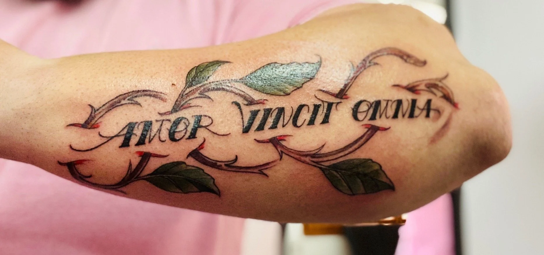 Tatuaje en el brazo con la frase "Amor siempre alma" rodeada de hojas y detalles decorativos en colores verde, negro y rojo.