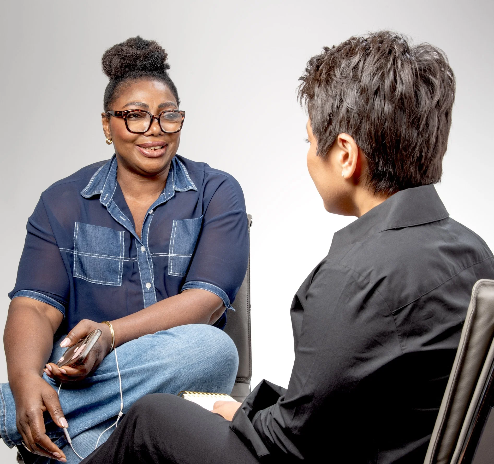 Two women having a conversation in a professional setting, one with glasses and a denim shirt, the other with short dark hair and a black shirt.