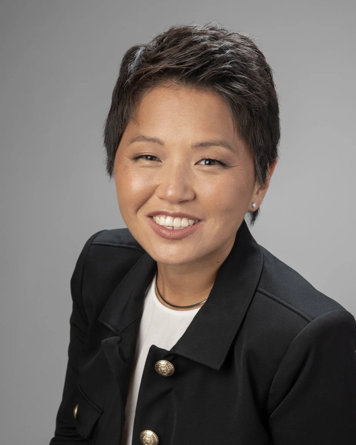 A woman with short dark hair smiling, wearing a black jacket with gold buttons, a white top, and a black choker, against a gray background.