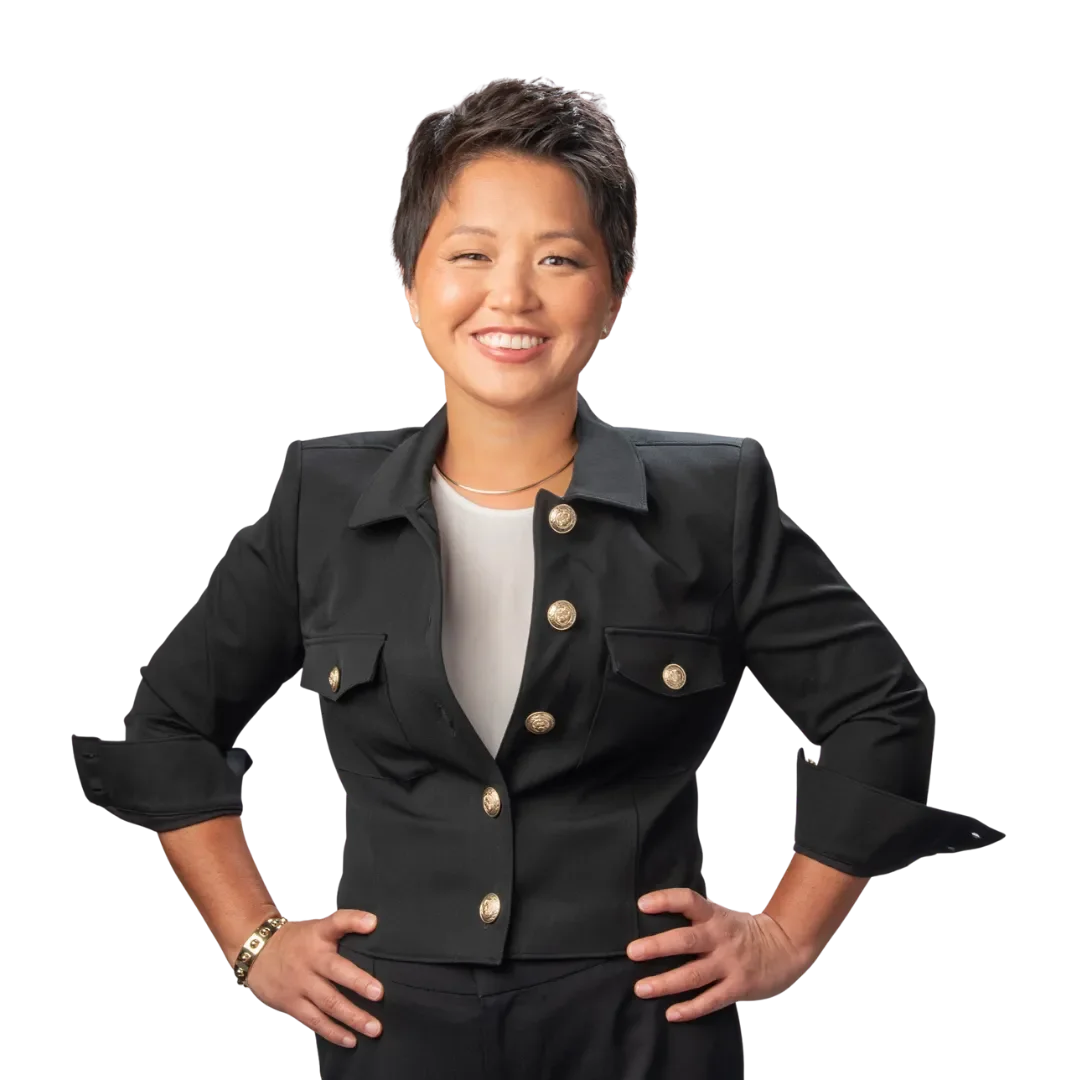 A woman with short hair smiling, wearing a black jacket with gold buttons and a white top, standing with her hands on her hips against a transparent background.