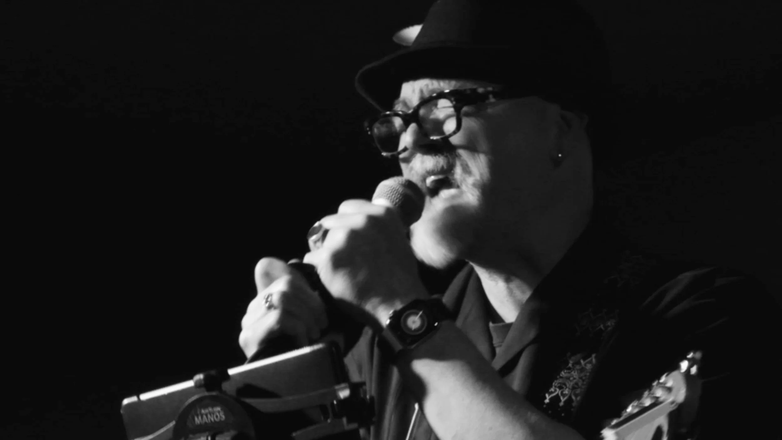A person wearing sunglasses and a hat, speaking into a microphone in a dark setting.