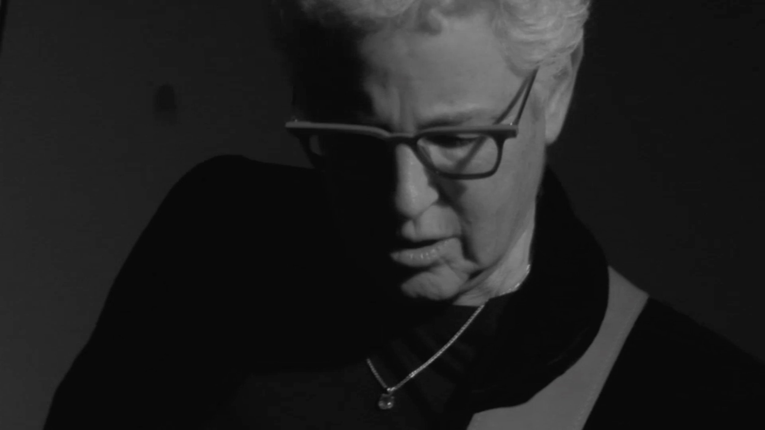 Black and white close-up of a woman wearing glasses, with short light-colored hair, looking down and slightly to the side.