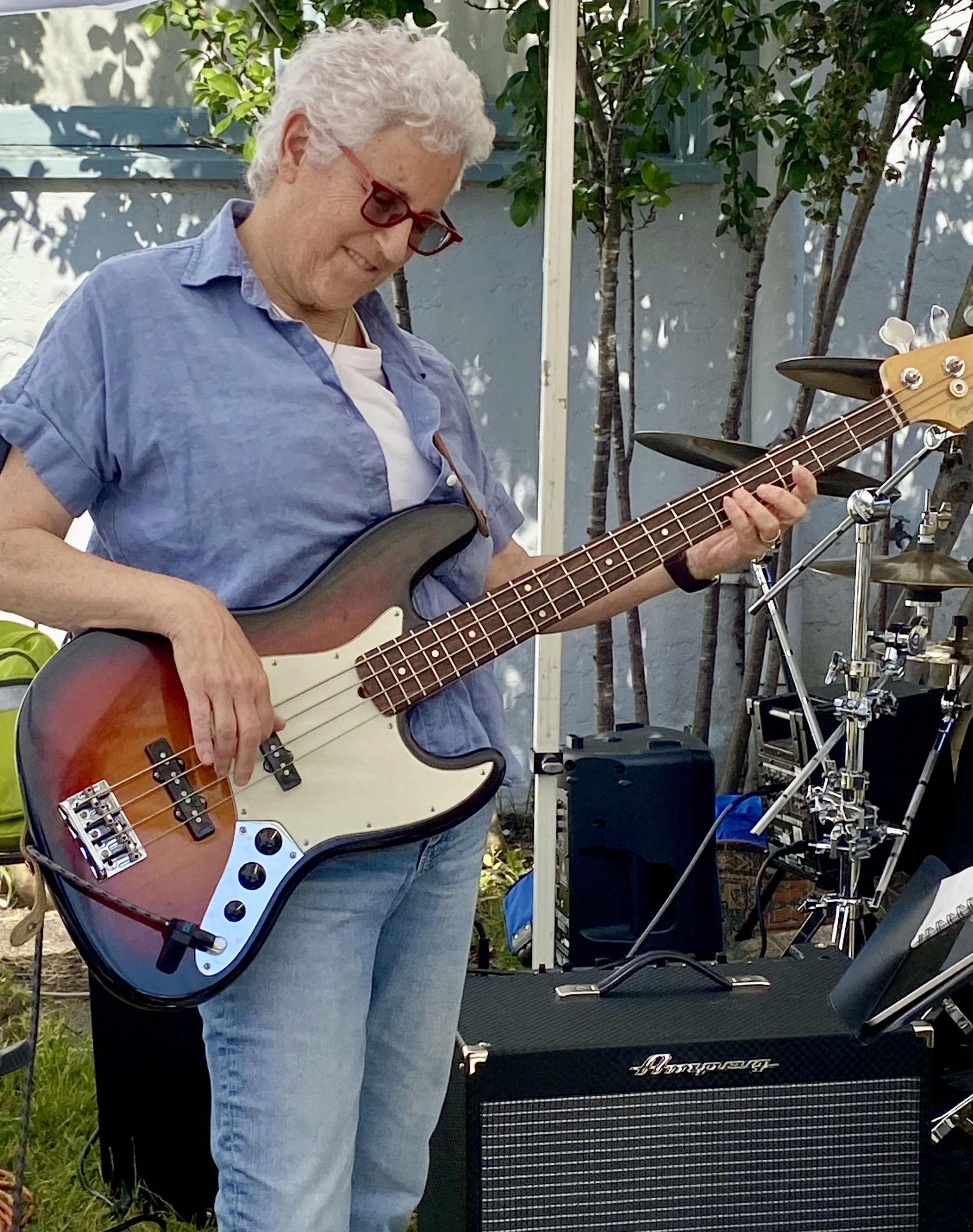Older woman with curly white hair and glasses playing an electric bass guitar outdoors, surrounded by trees and musical equipment, including an amplifier and drum set.