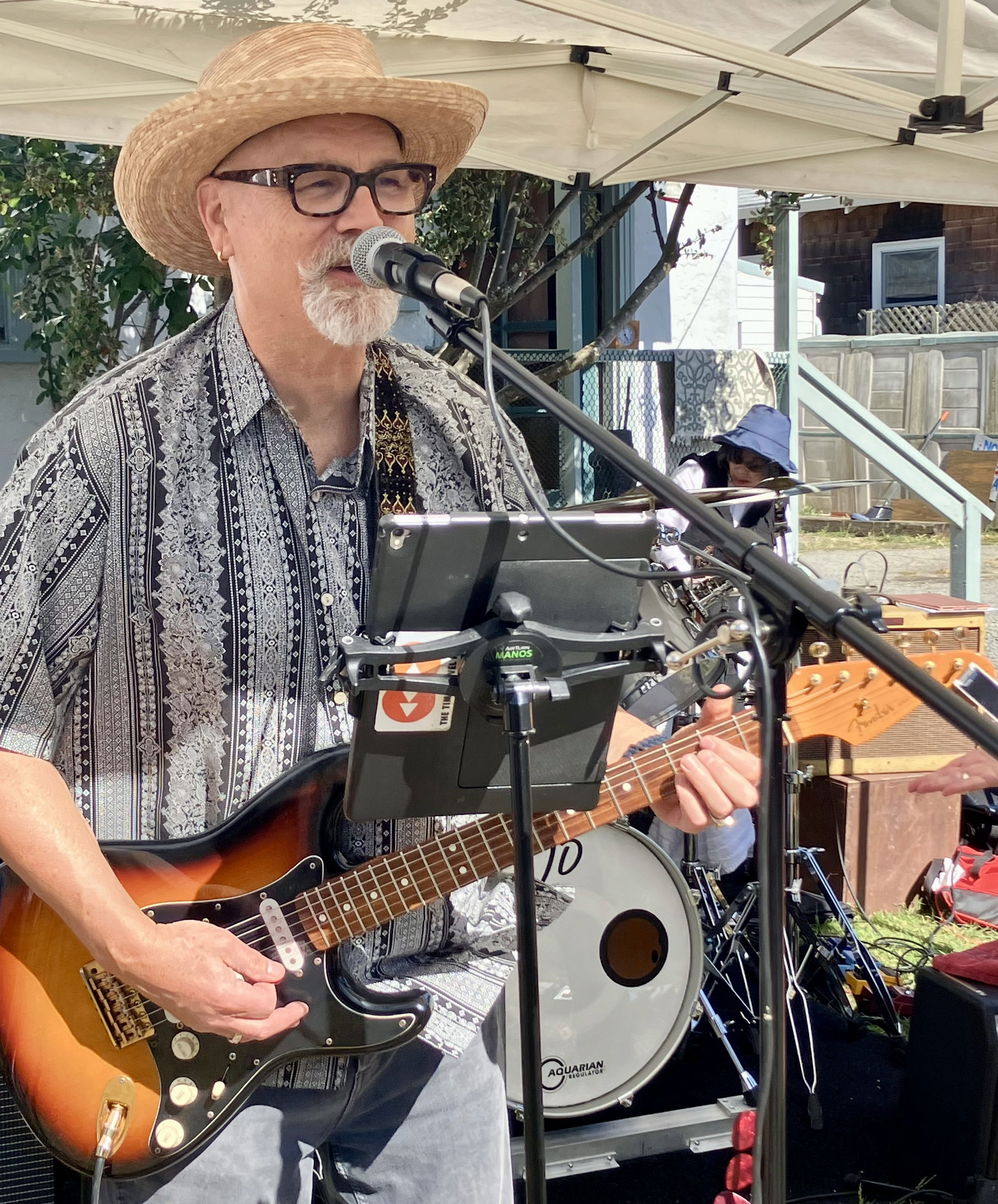 A man wearing a straw hat, glasses, and a patterned shirt is singing into a microphone while playing an electric guitar during an outdoor performance. In the background, there is a drum set and another person wearing a blue hat playing a keyboard, under a white canopy with trees and residential houses in the surroundings.