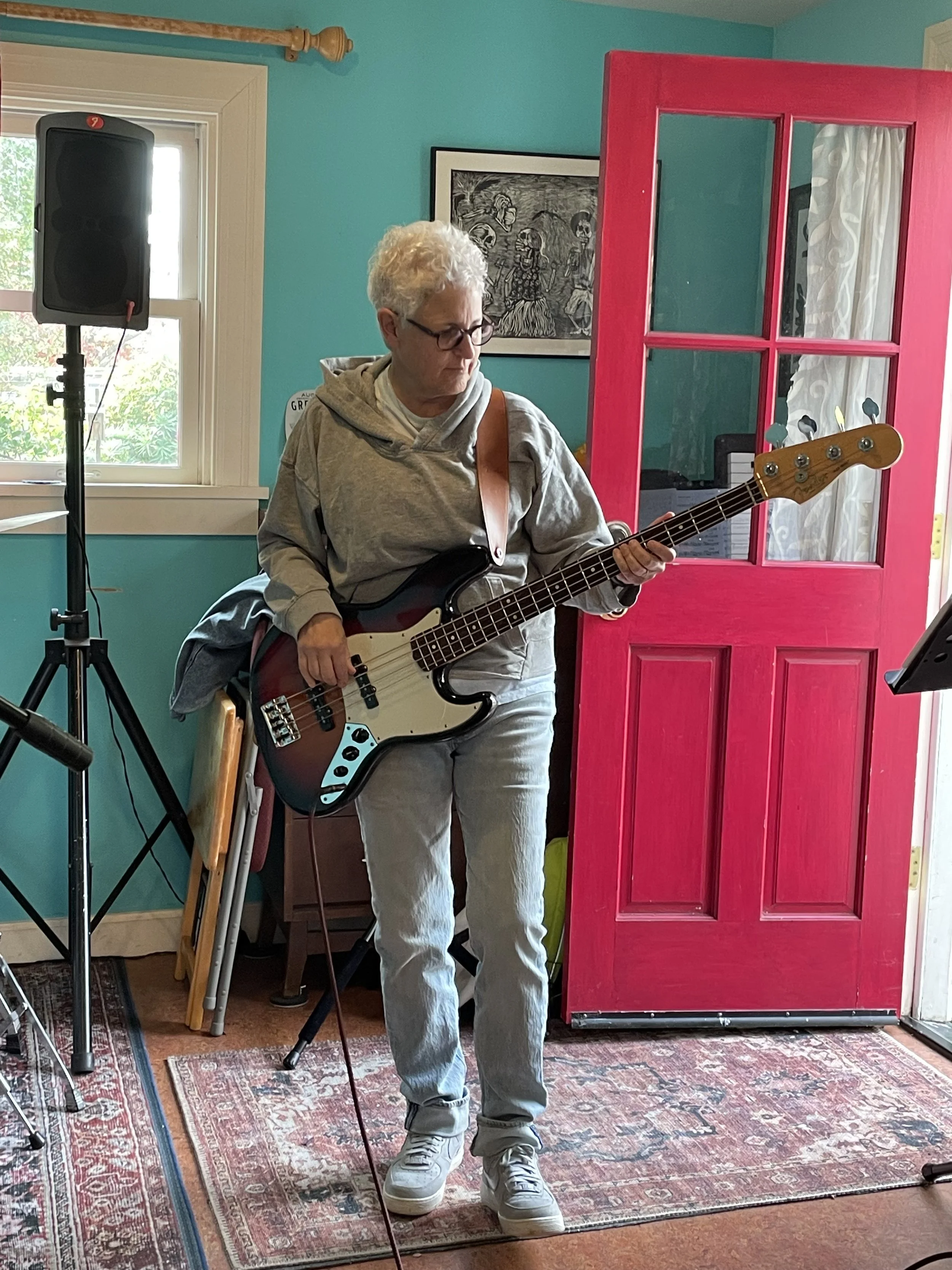 An elderly woman playing an electric bass guitar indoors, standing on a vintage rug near a bright pink door and blue wall, with sound equipment nearby.