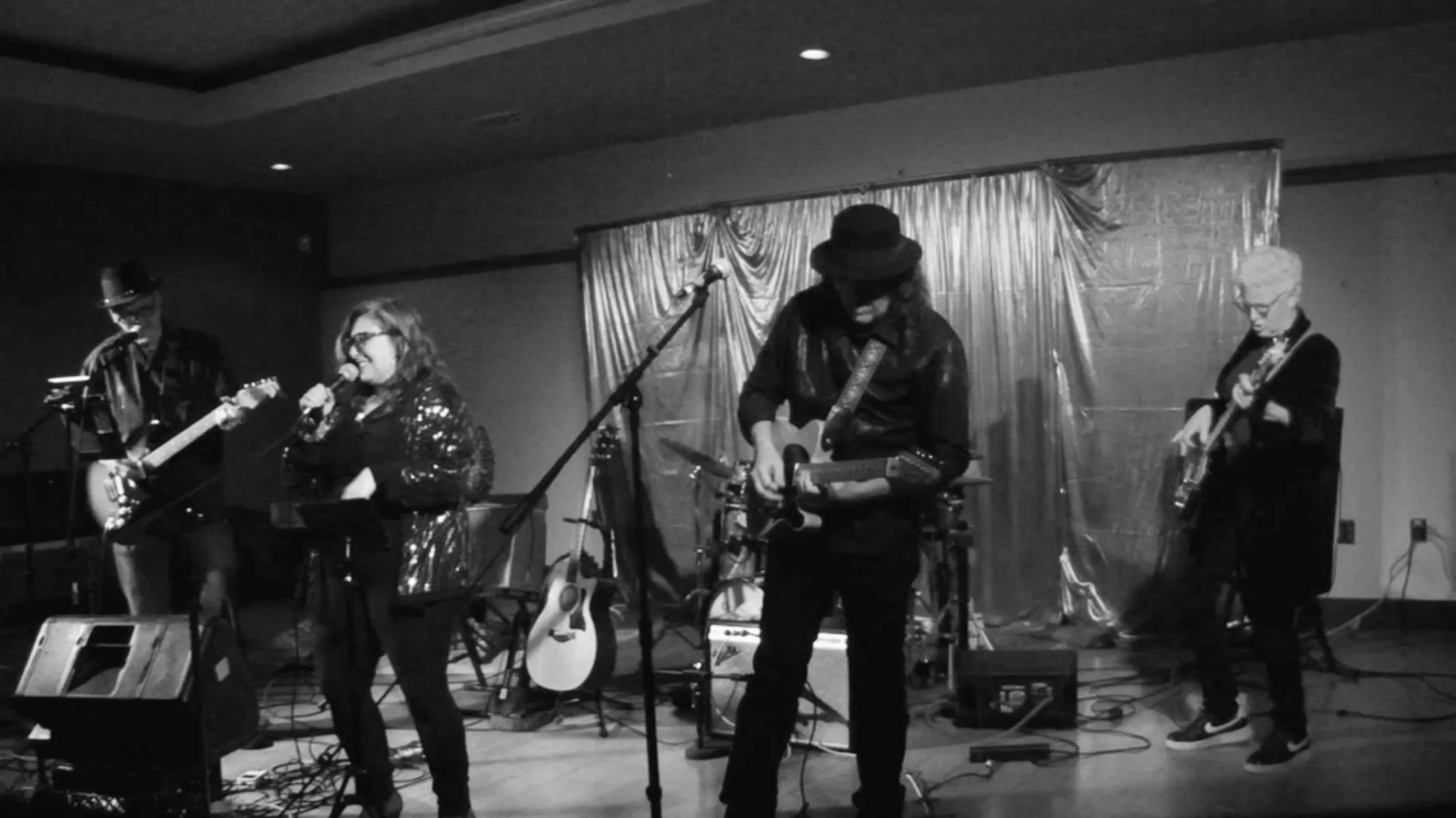 A black and white photo of a live music performance featuring four musicians on stage with guitars and microphones, set against a curtain backdrop.
