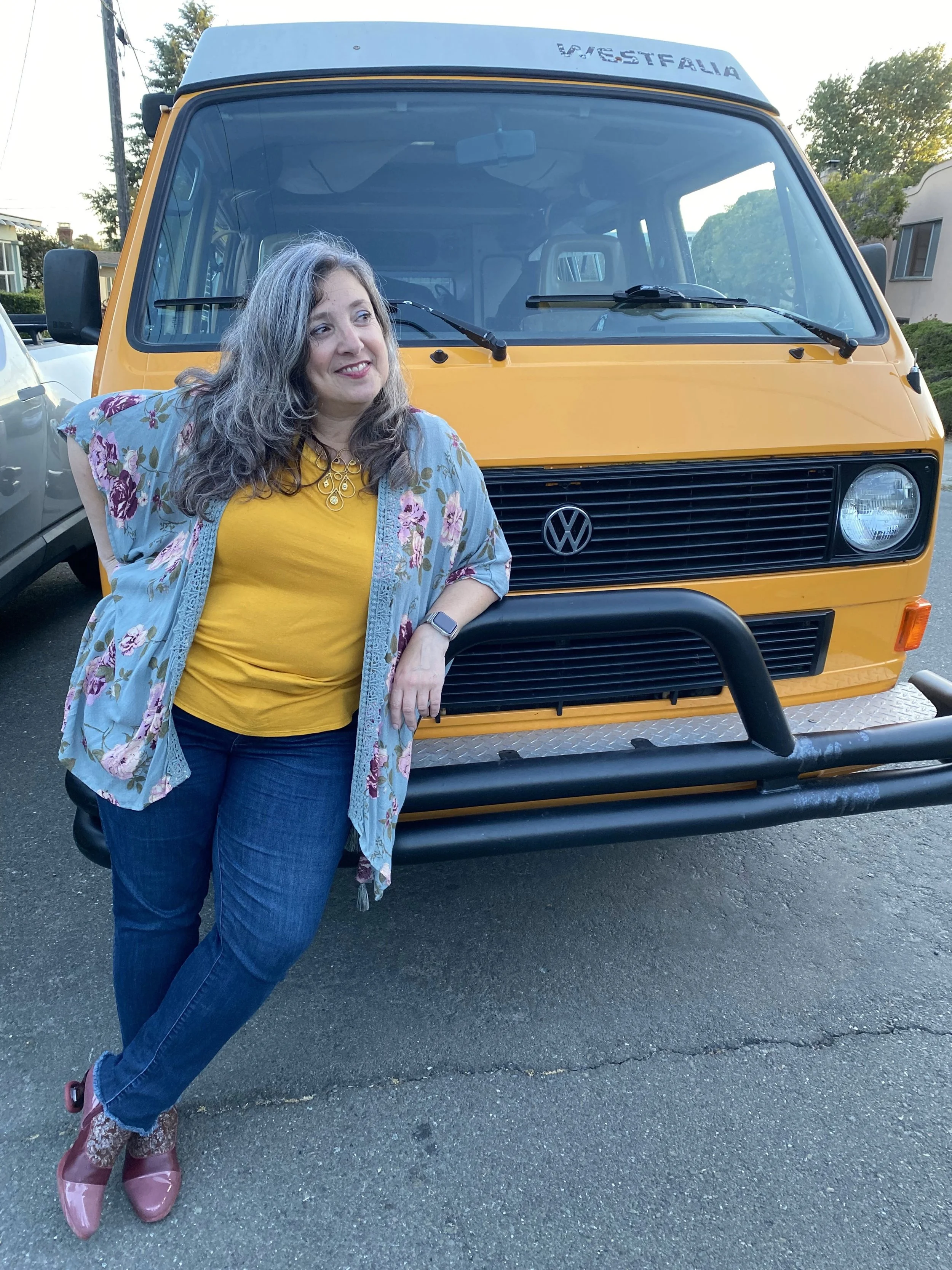 A woman with curly gray hair wearing a yellow shirt, blue jeans, a floral open jacket, and pink rain boots standing in front of a yellow Volkswagen Westfalia camper van on a paved street during daytime.