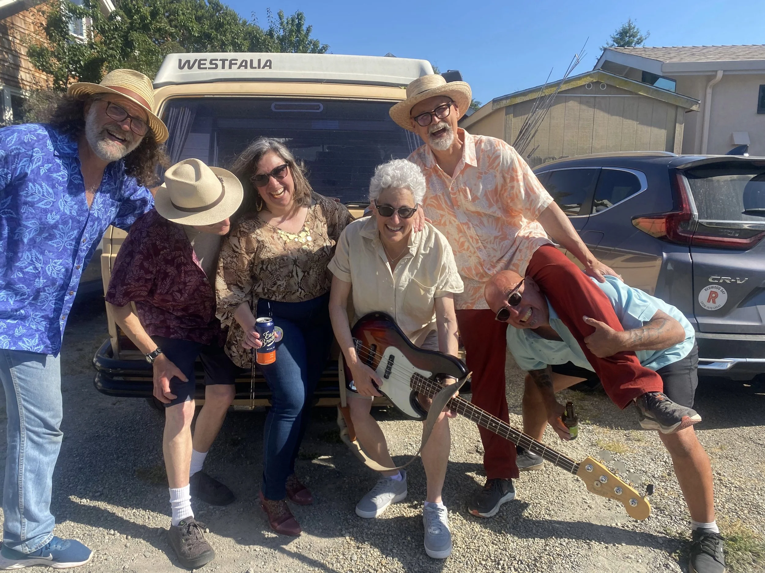 A group of seven older adults smiling and posing outdoors in front of a vintage camper and a car on a sunny day. They are wearing casual summer clothing and hats, some holding drinks, with one person playing a guitar.