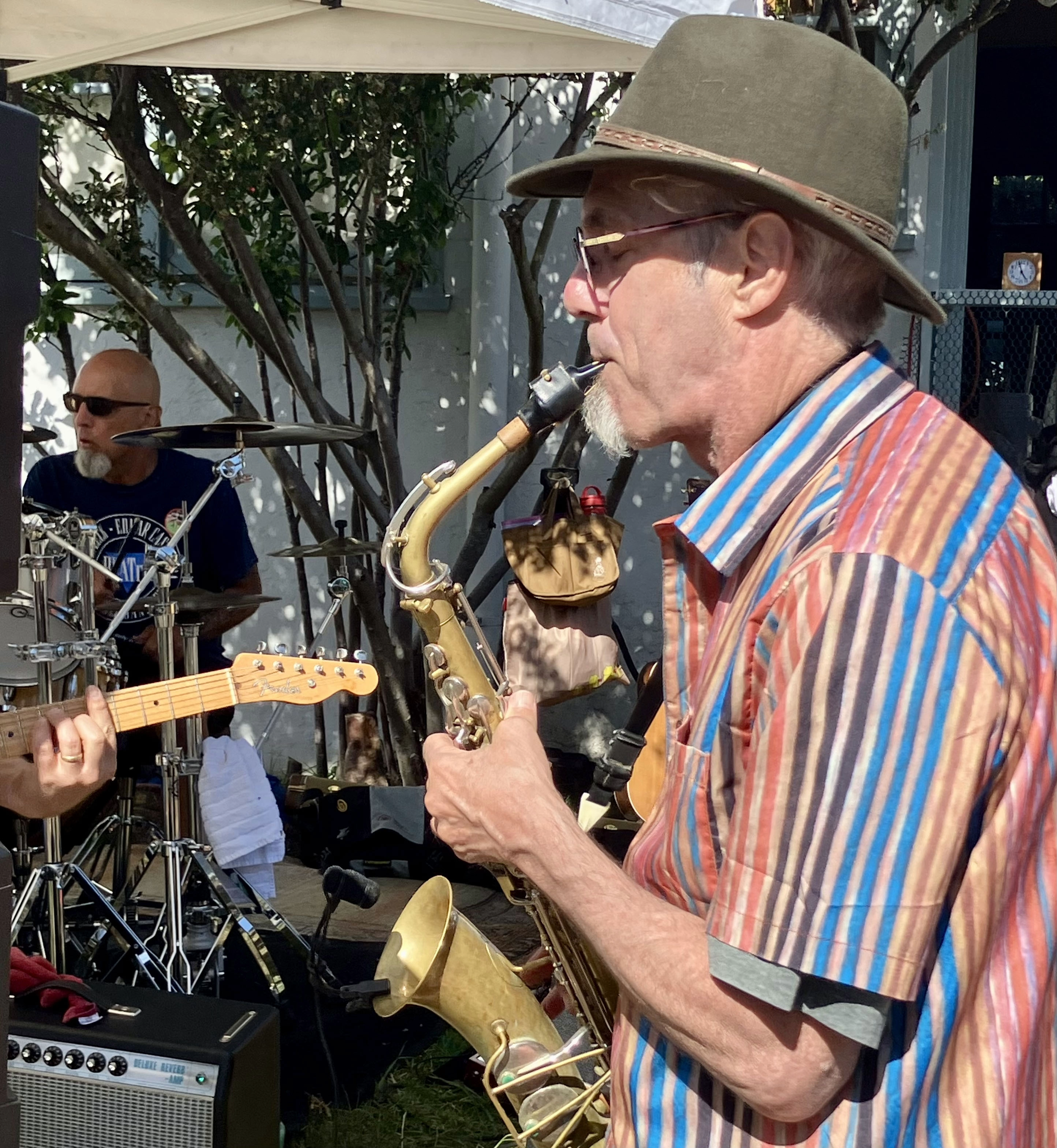 A man wearing a wide-brimmed hat and sunglasses playing a saxophone at an outdoor event. Another man with a beard and sunglasses is playing drums in the background. There are trees, a tent, and musical equipment including an amplifier in the scene.