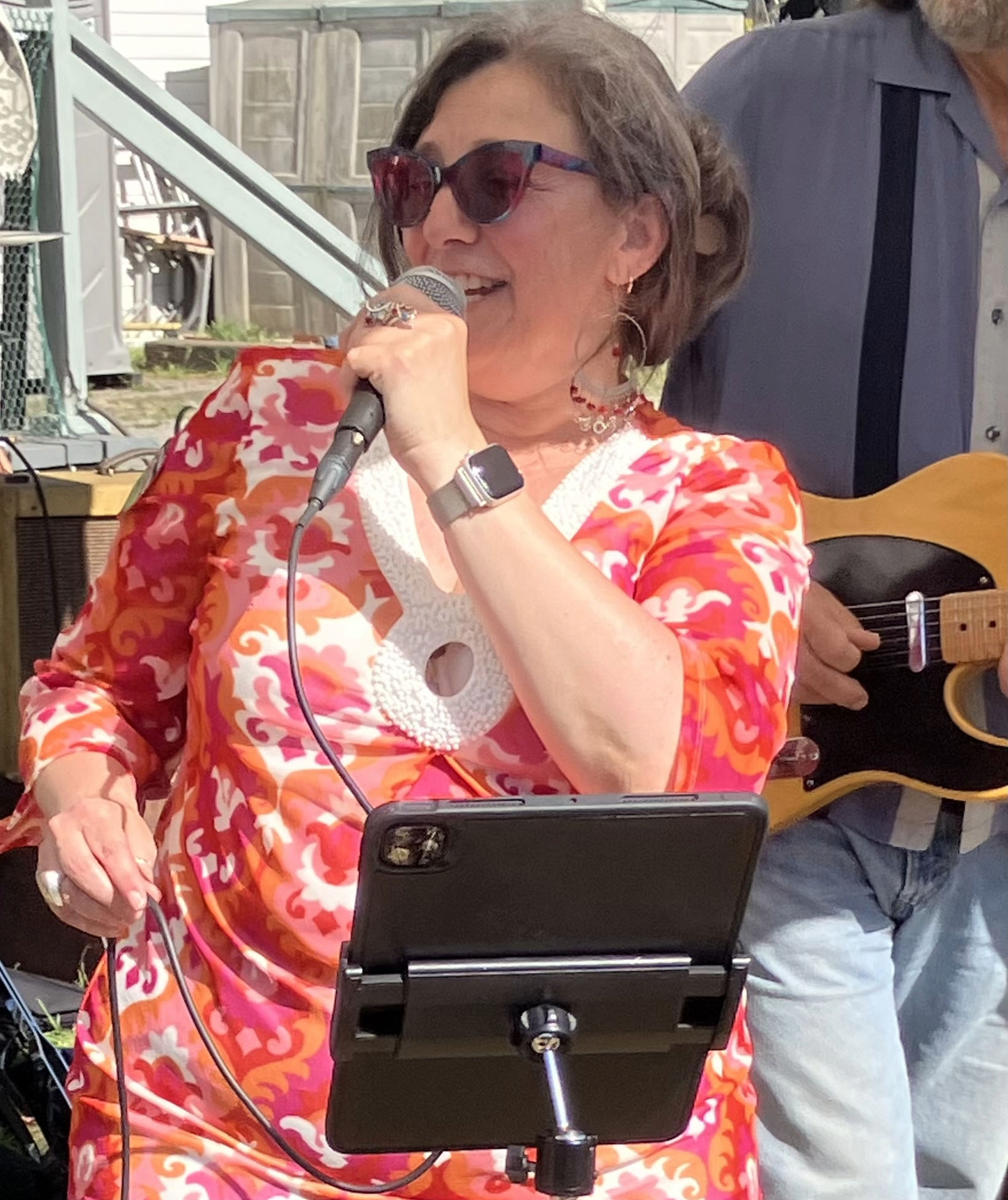 A woman wearing sunglasses and colorful dress singing into a microphone, standing outdoors with a music stand in front of her.