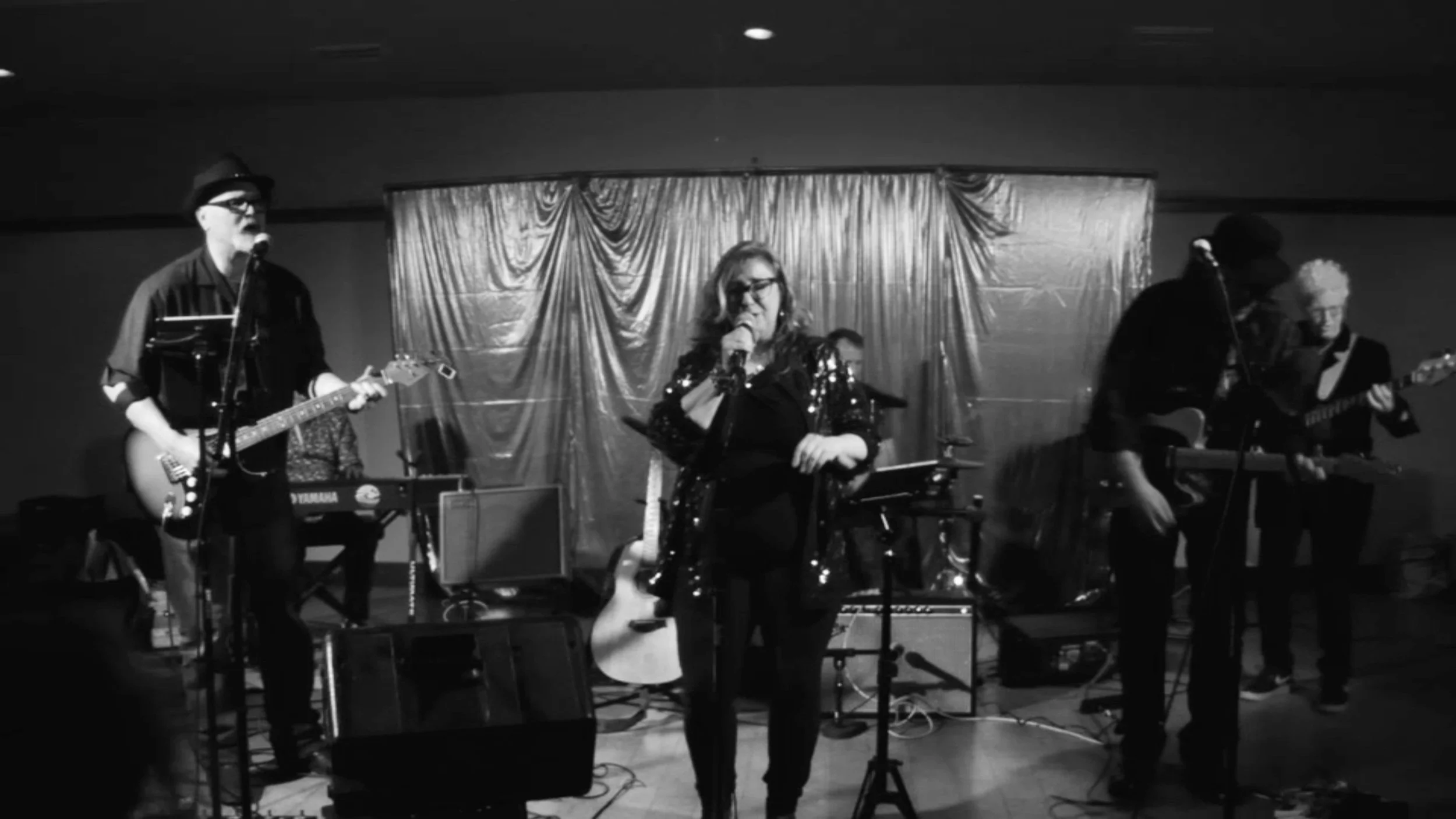 A band performing on stage with three musicians playing guitars and a woman singing into a microphone, with a curtain backdrop.