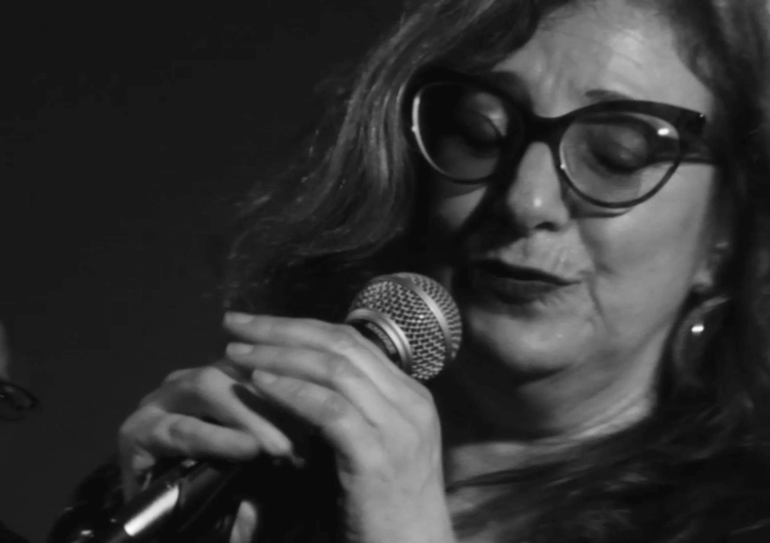 A black and white close-up of a woman with curly hair and glasses, singing or speaking into a microphone.