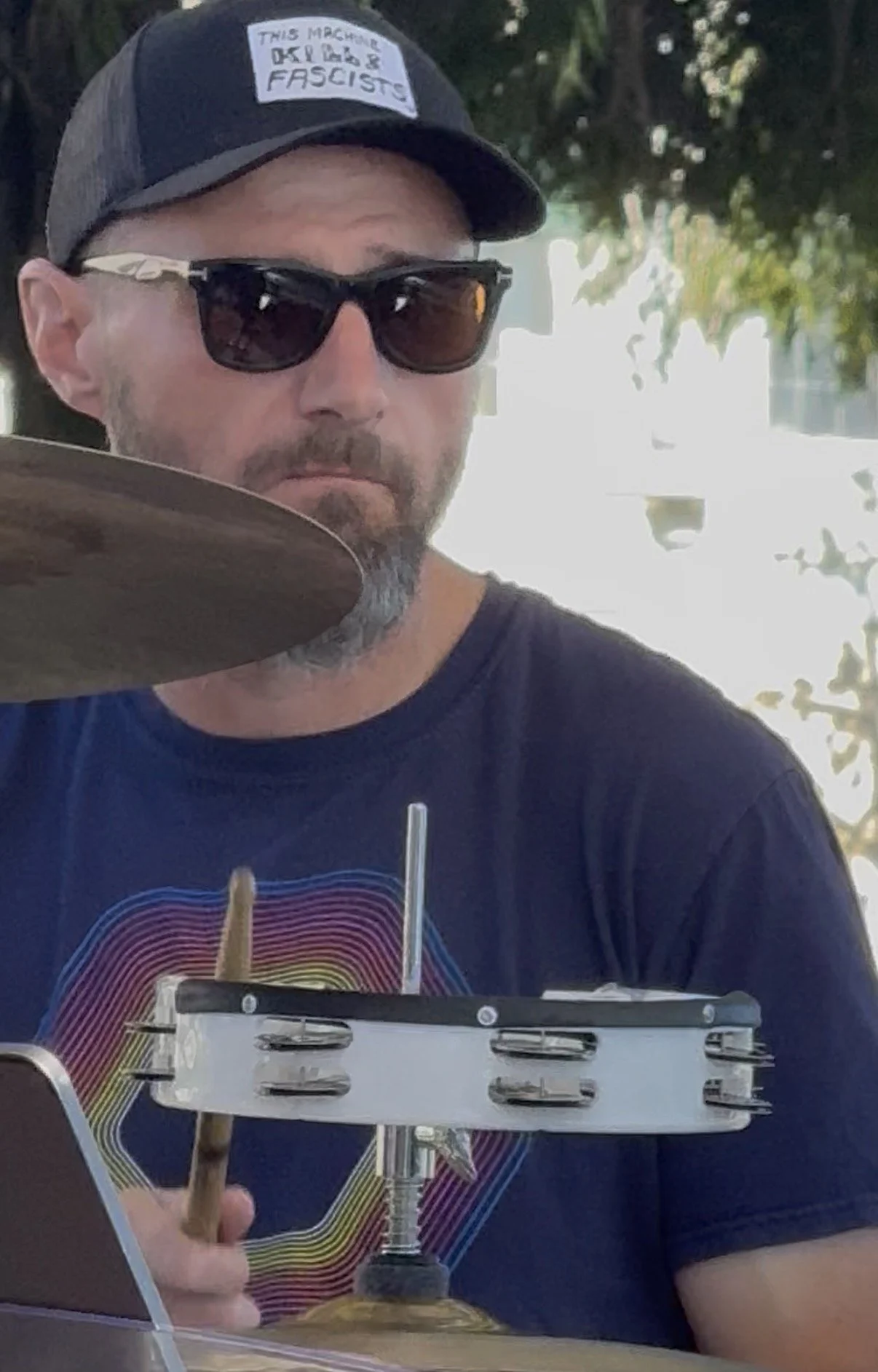 A man wearing sunglasses and a black baseball cap with a sticker reading 'This machine is operated by fascists' is sitting outdoors. He is holding a drumstick and playing a drum pad, with a drink in front of him and blurred trees in the background.