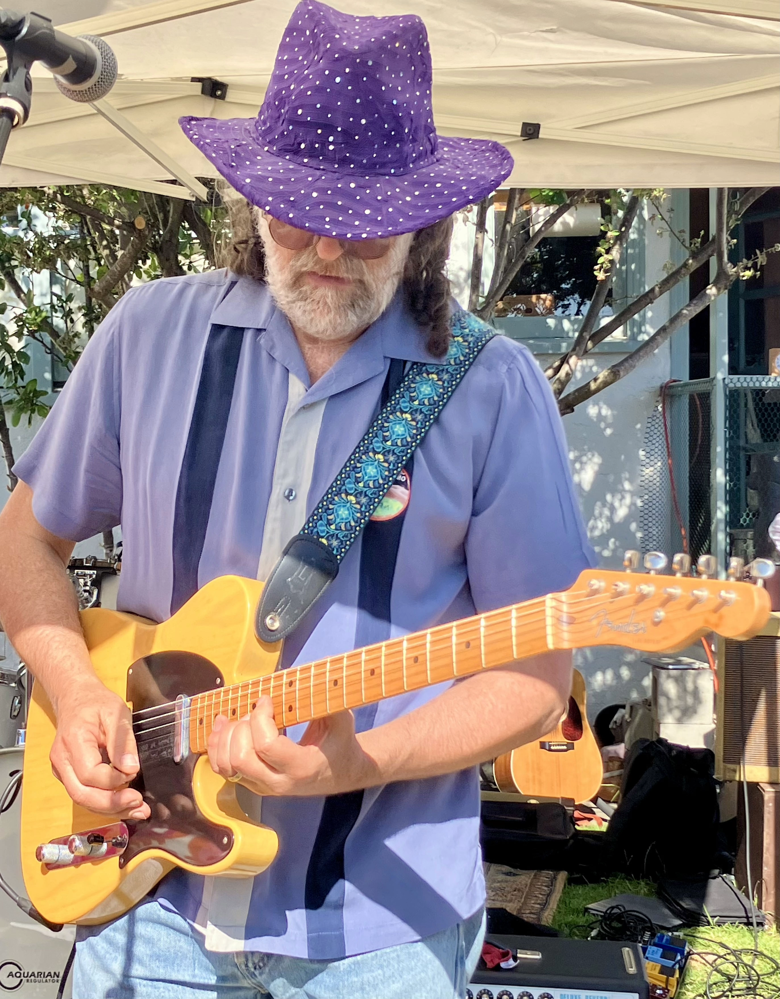 A man wearing a purple polka-dot wide-brimmed hat, sunglasses, and a blue button-up shirt, playing an acoustic guitar at an outdoor event surrounded by equipment and trees.
