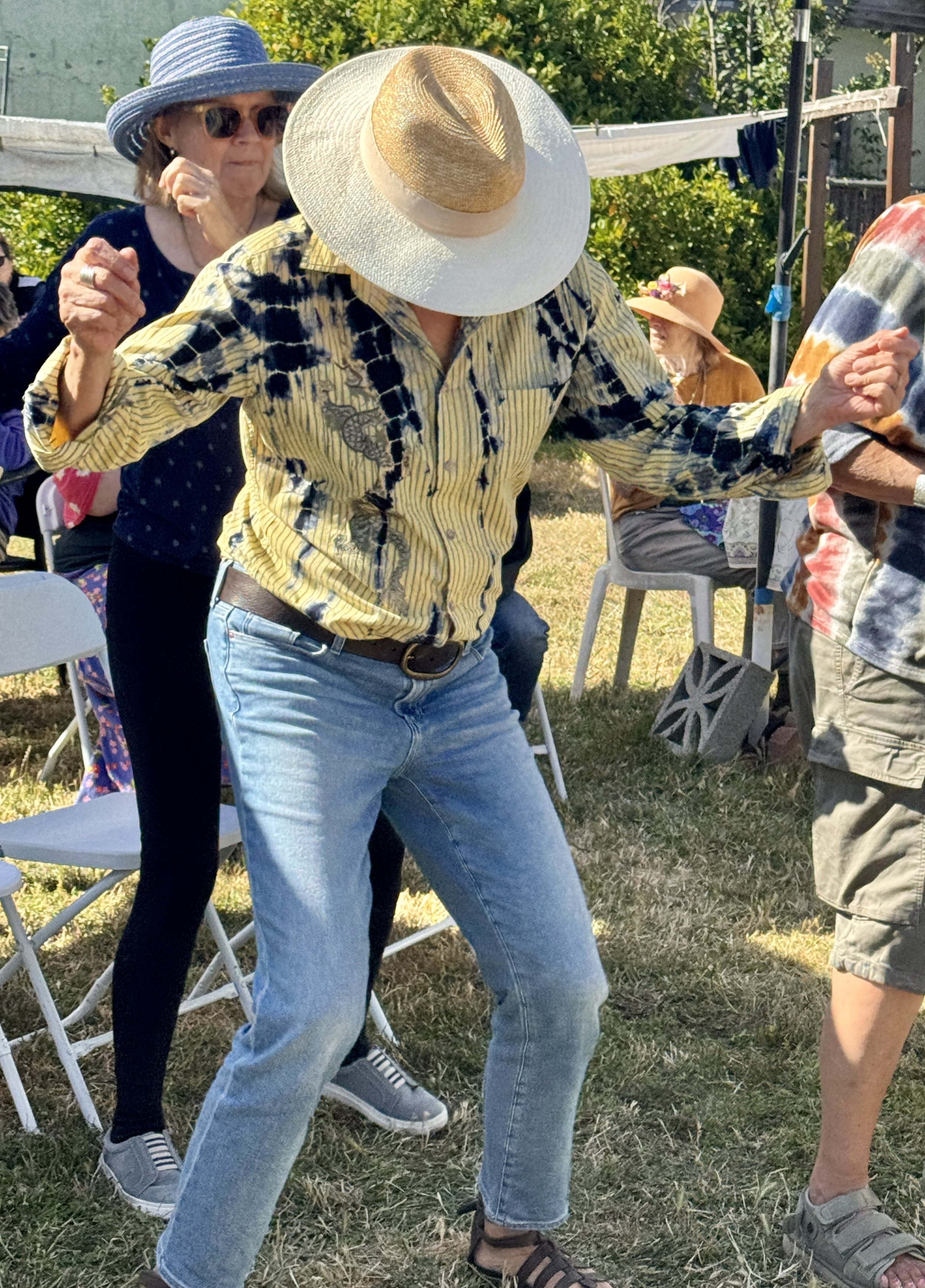 People dancing outdoors on grass, wearing summer clothing and hats, with some sitting on chairs in the background.