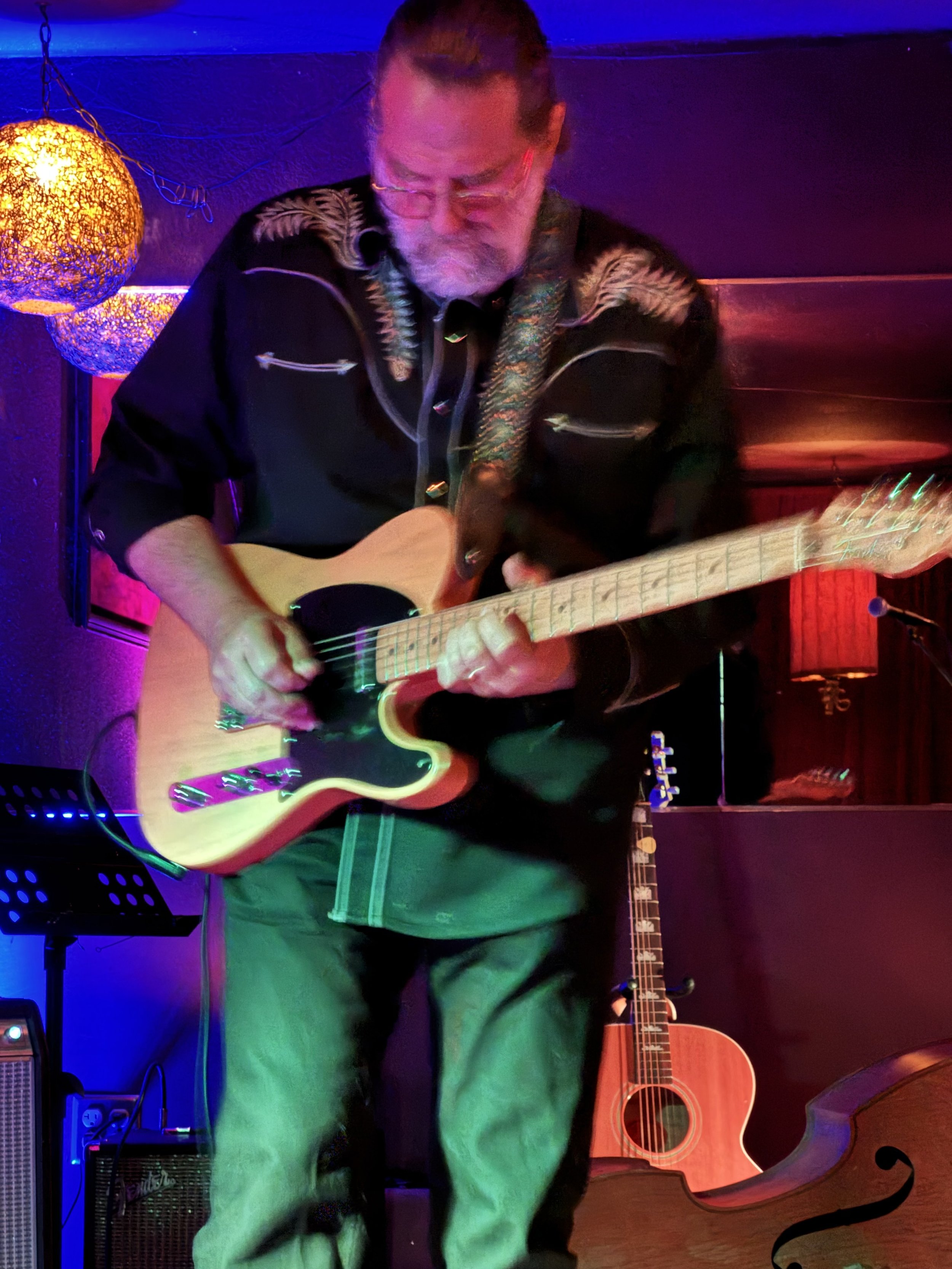 A man with glasses and a beard playing an acoustic guitar on a stage with colorful lights and a large wooden double bass in the background.