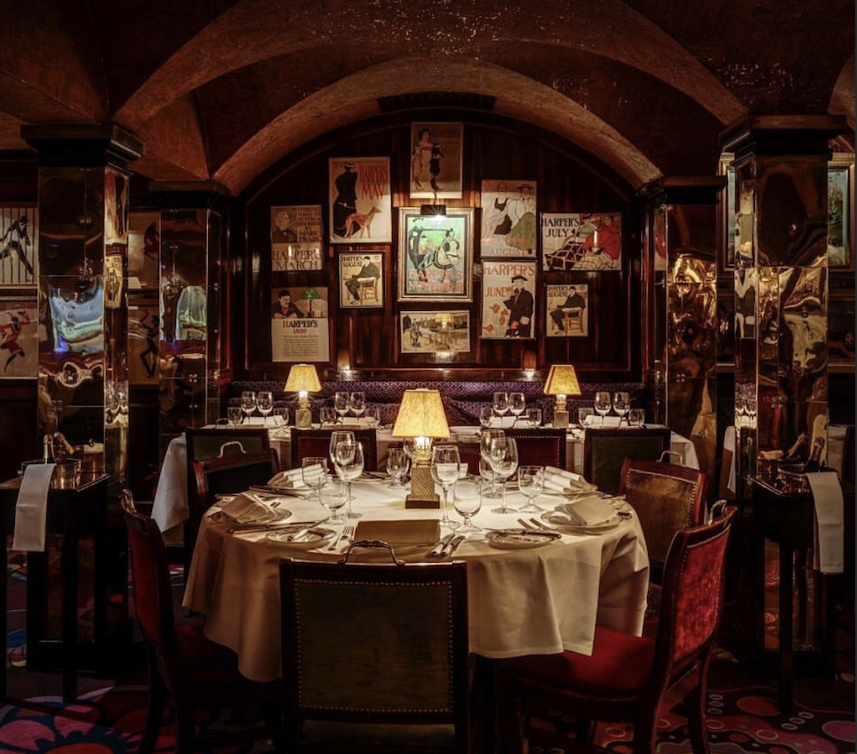 Elegant restaurant dining room with round table set with white tablecloth, wine glasses, and cutlery, illuminated by small lamps, with framed vintage posters on dark wood-paneled walls and arched ceiling.