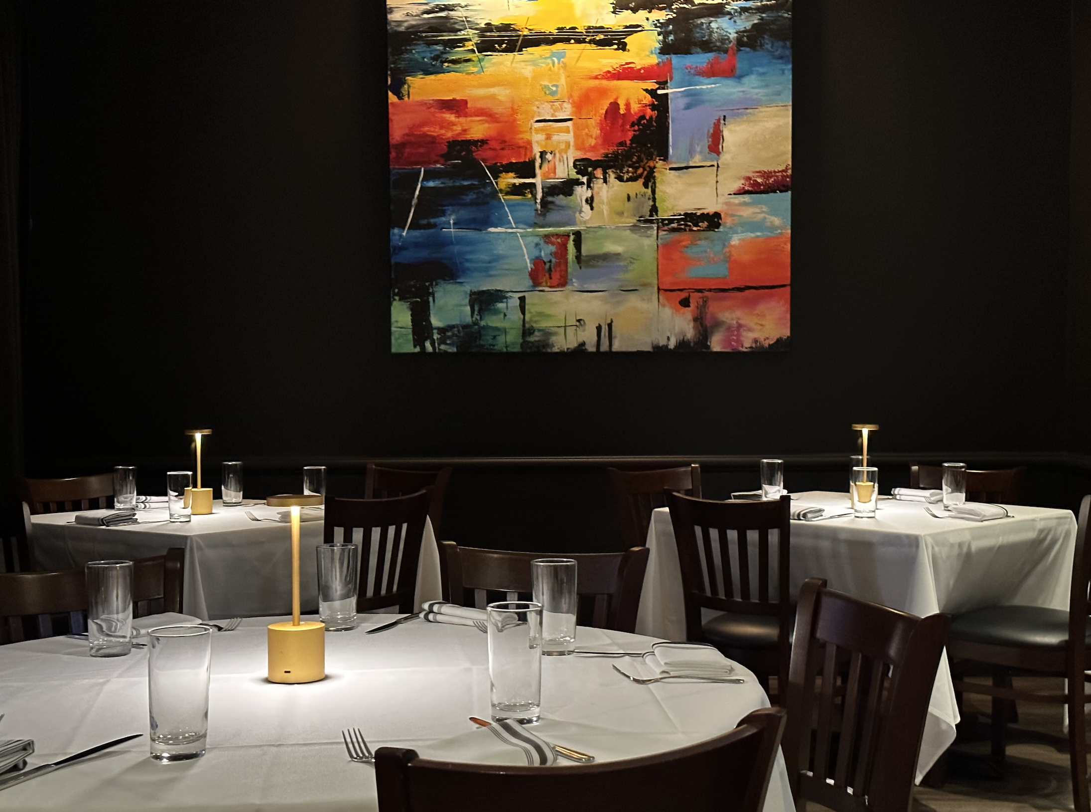 Elegant restaurant interior with white tablecloths, wooden chairs, and modern yellow table lamps. A colorful abstract painting hangs on the dark wall.