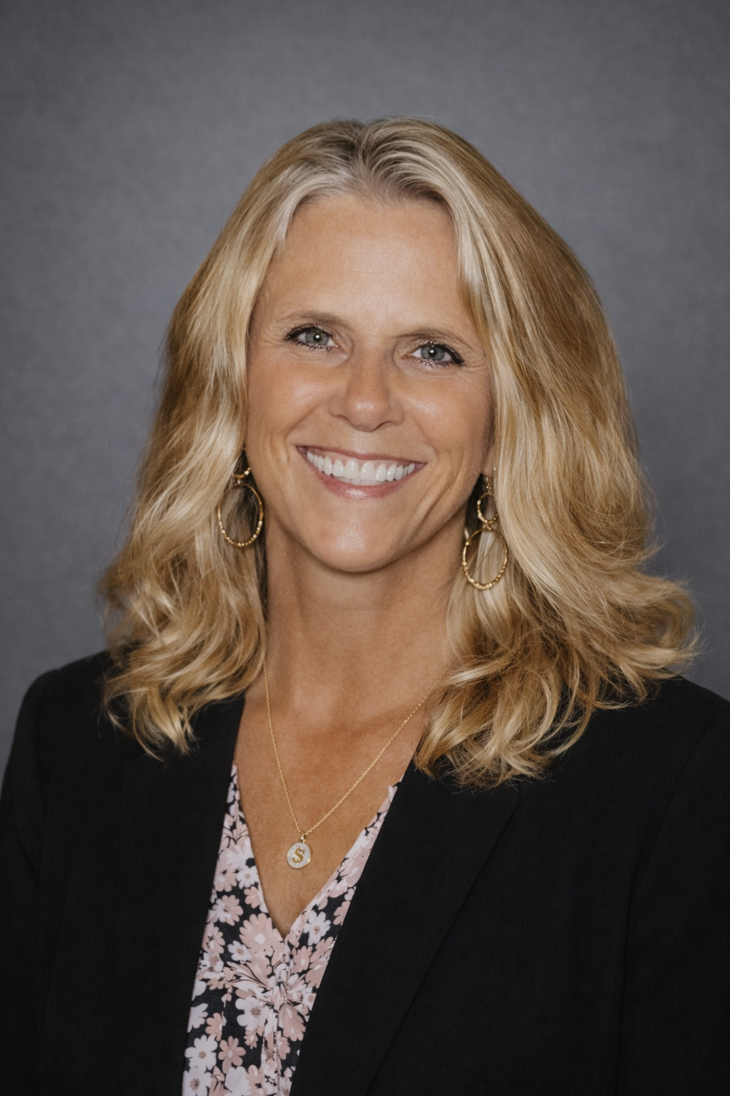 A woman with blonde, wavy hair wearing a black blazer, floral blouse, gold hoop earrings, and a gold necklace with a dollar sign pendant, smiling against a gray background.