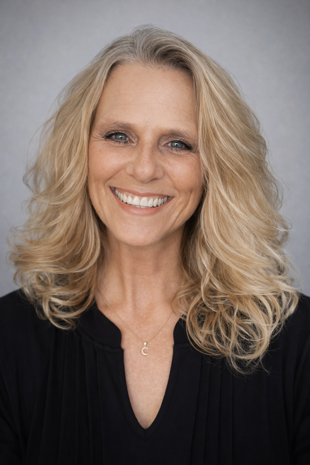 A smiling woman with blonde, curly hair wearing a black top and a gold necklace with a letter 'C' pendant.