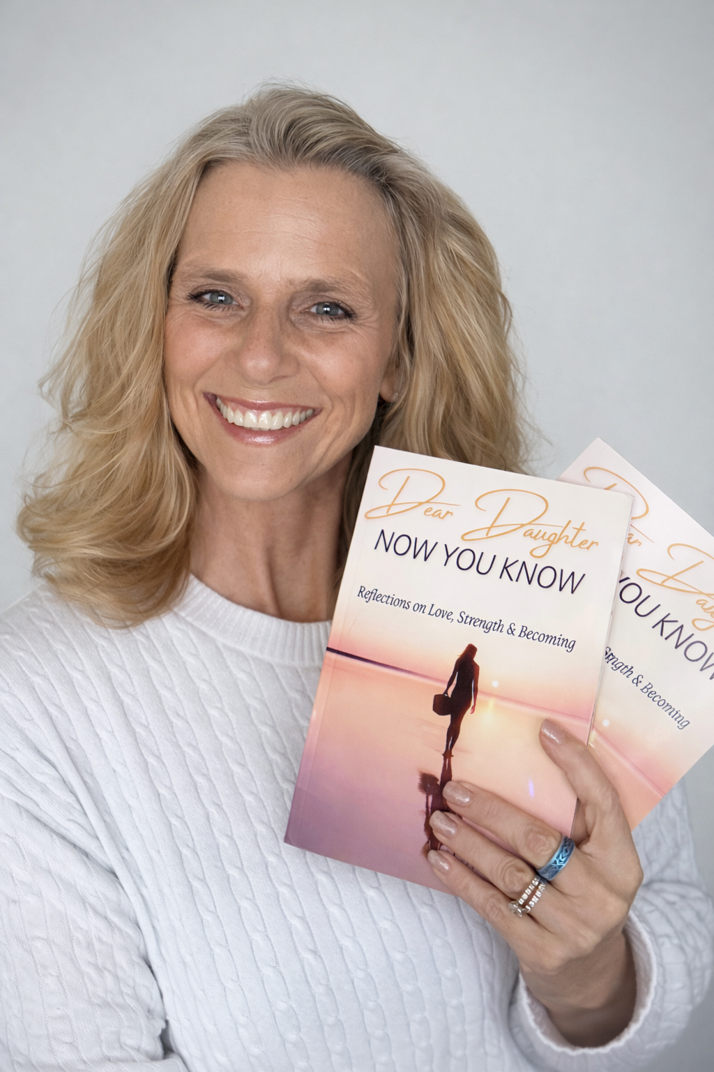 A smiling woman holding a book titled "Dear Daughter, Now You Know: Reflections on Love, Strength & Becoming".