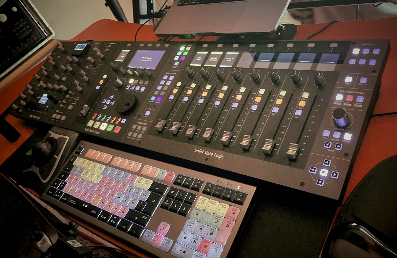 Table de mixage audio professionnelle avec plusieurs boutons, faders et écrans, accompagnée d'un contrôleur de musique avec des touches colorées, sur une table en bois.