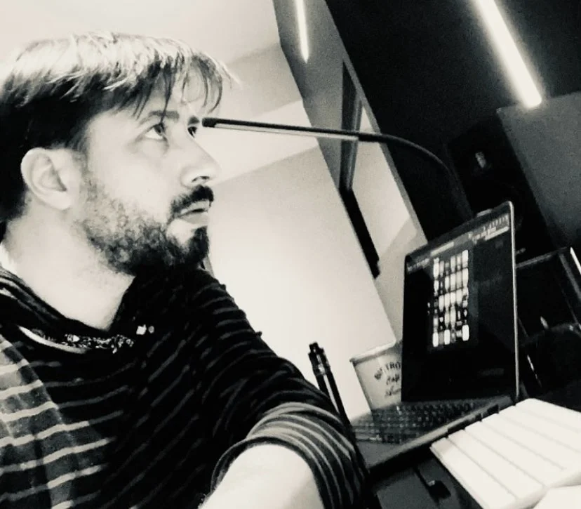 Un homme avec des cheveux bruns, une barbe, portant un t-shirt à rayures, travaille sur un ordinateur portable dans un environnement de bureau ou studio, avec une tasse à côté.