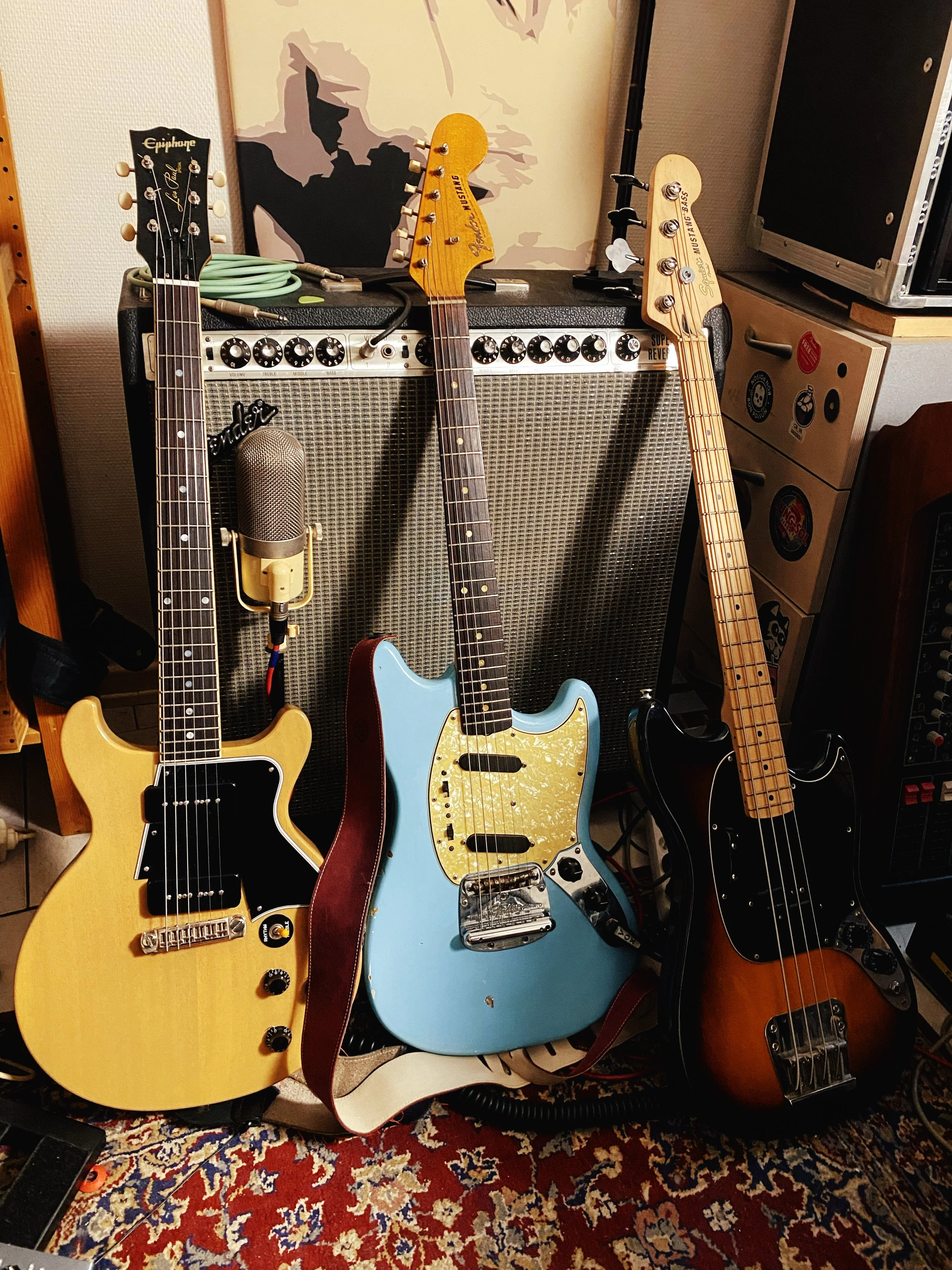 Trois guitares électriques posées devant un ampli à lampe, avec un microphone suspendu devant ; décoration artistique en arrière-plan.
