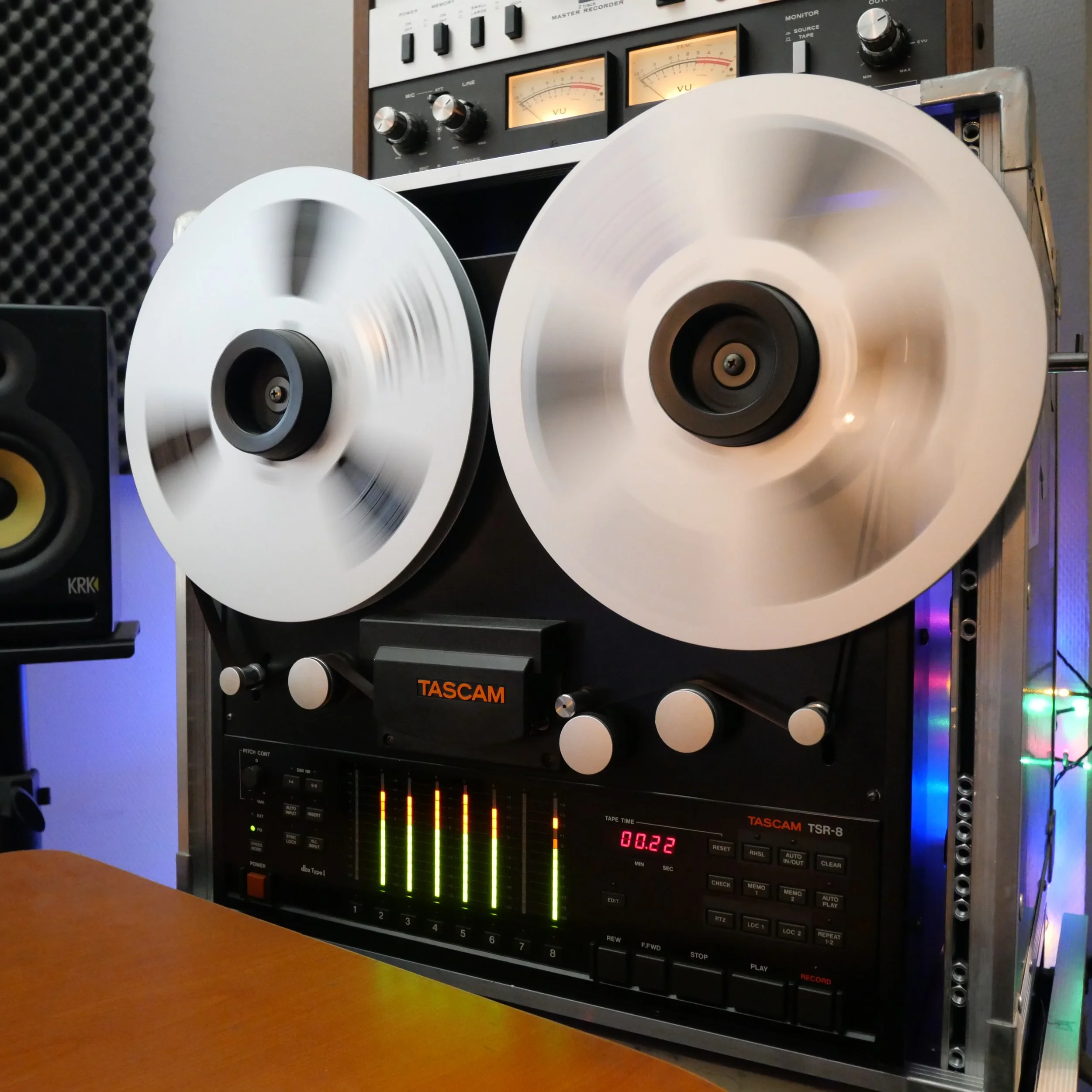 Enregistreur Tascam TSR-8, un vieux magnétophone à bobines, avec deux bobines blanches en mouvement, dans un studio avec des panneaux acoustiques.