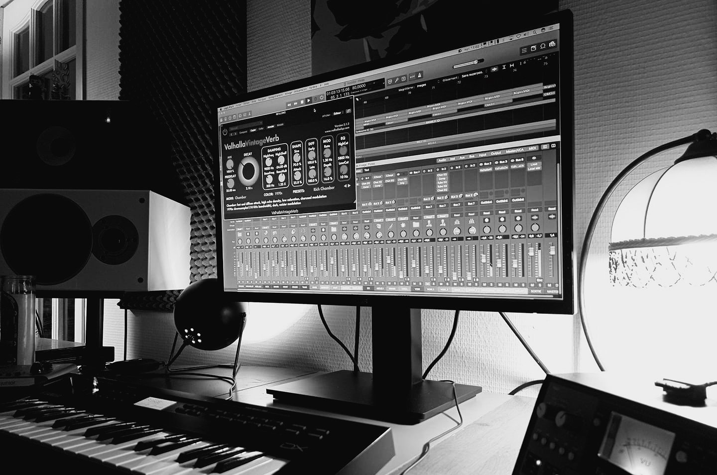 Studio de musique avec un écran d'ordinateur affichant une station de travail audio numérique, haut-parleurs, clavier de piano, lampadaire, et éléments acoustiques. Enregistrement ou production musicale en cours.