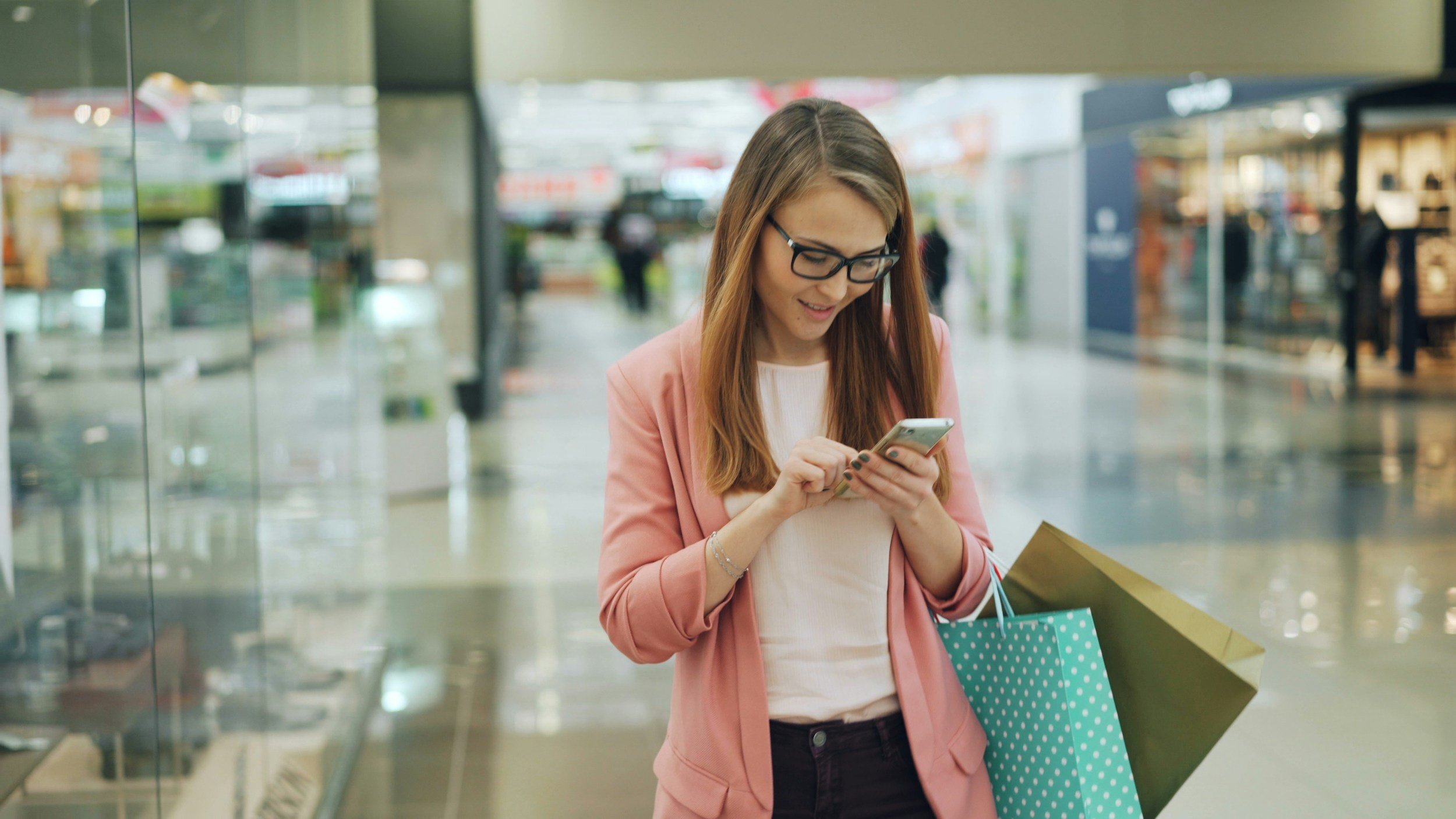 Young woman with glasses in pink blazer holding shopping bags and using her smartphone inside a shopping mall.