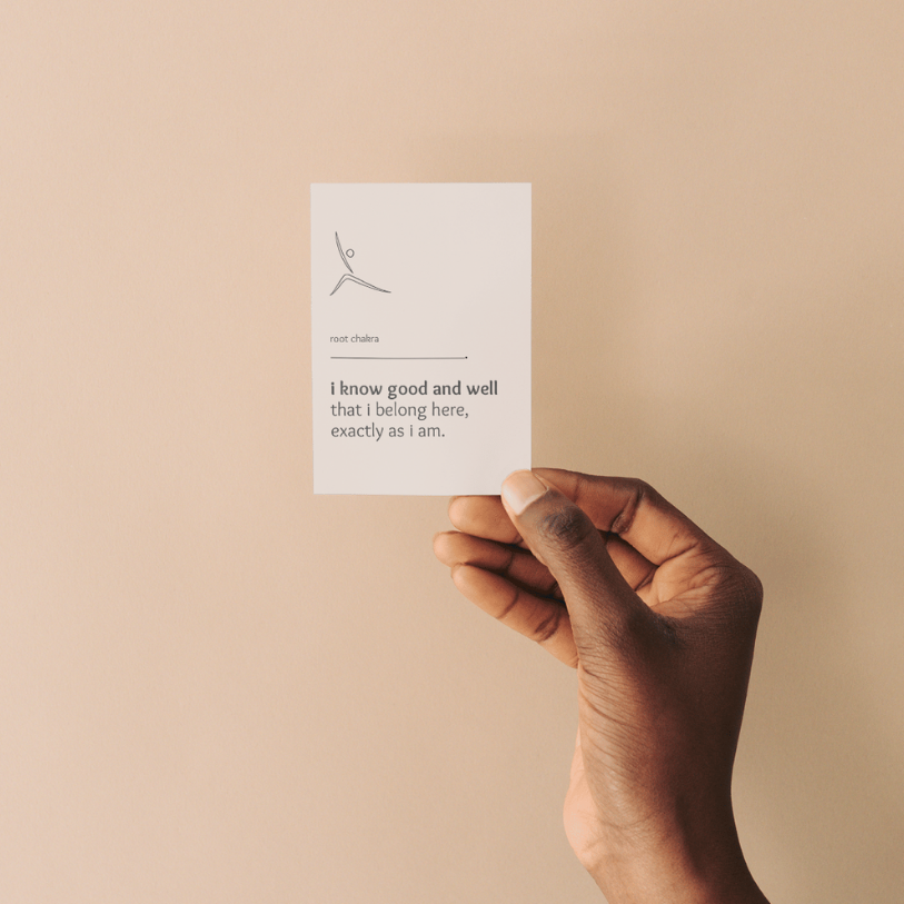 A person holding a white card with a minimalist illustration and text about root chakra against a beige wall.