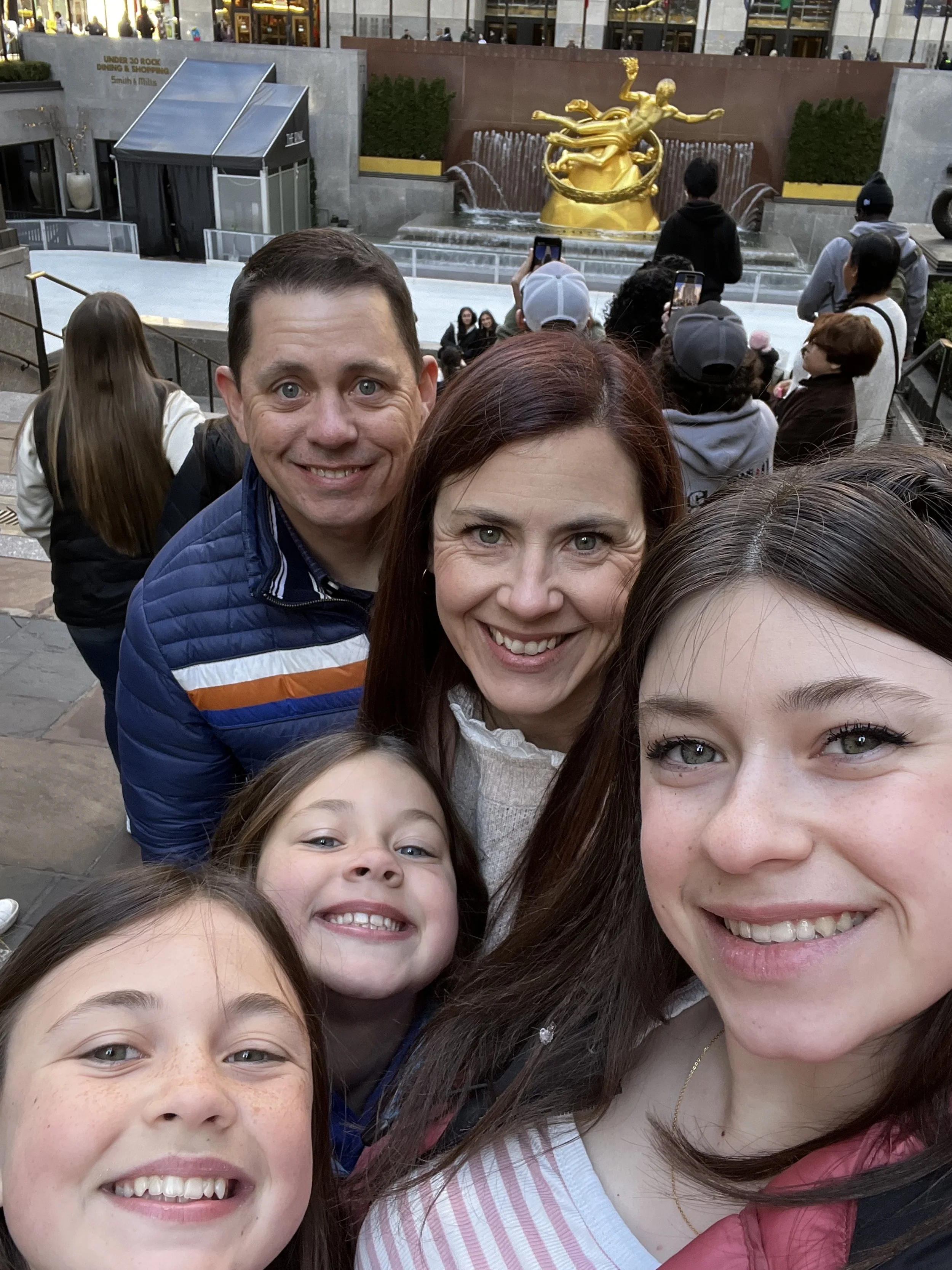 Family of five taking a selfie in front of a fountain with a golden sculpture, surrounded by other people.